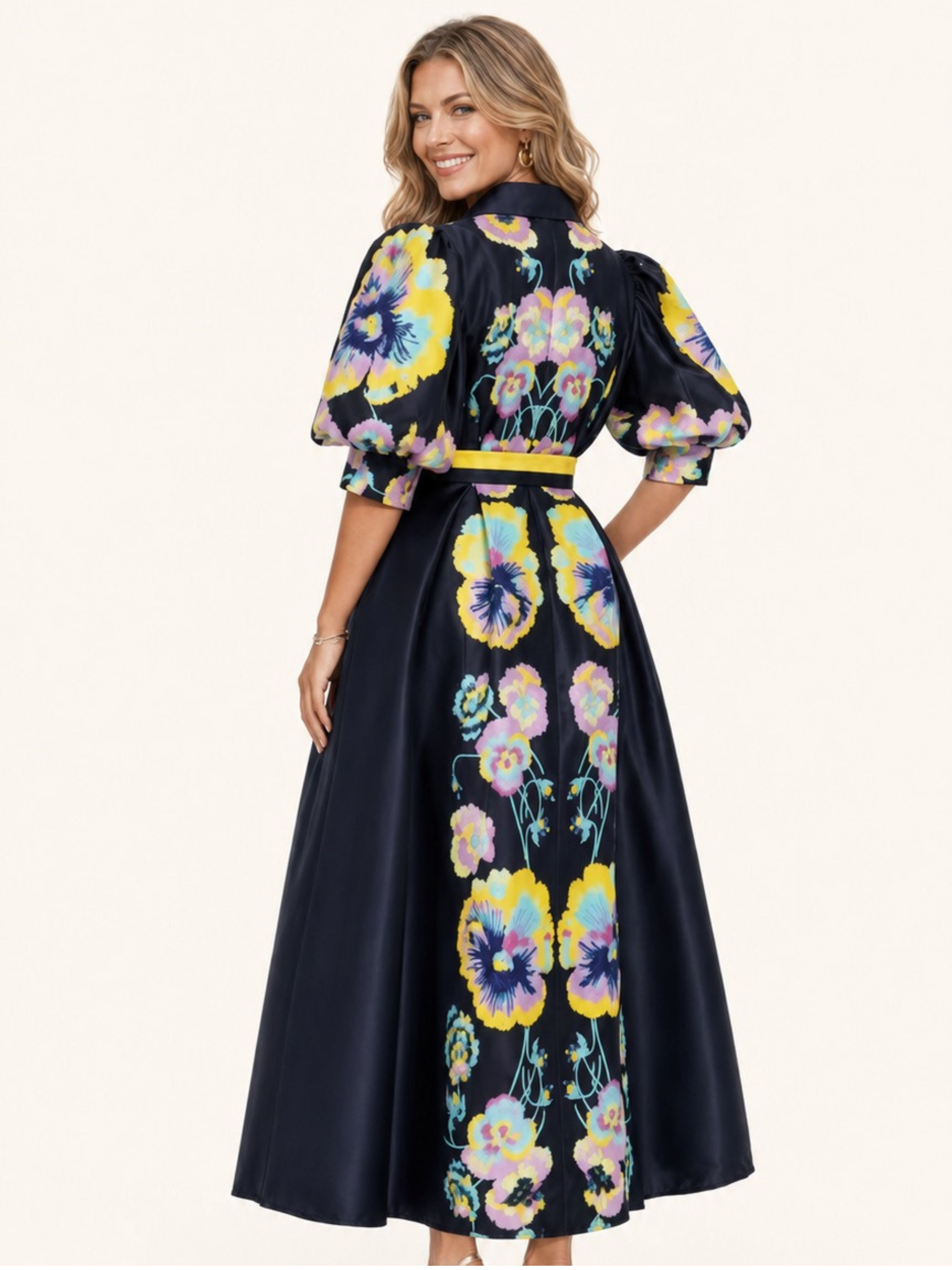 A woman modeling a navy blue maxi dress with a vibrant multicolor floral print, featuring puff sleeves and a matching waist tie, set against a plain off-white background.