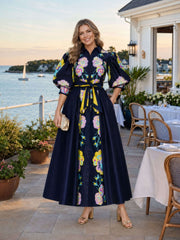 A woman stands on a seaside patio wearing a navy maxi dress with large pink, yellow, and white floral print, puff sleeves, and a matching waist tie. She is holding a small metallic clutch and wearing beige heeled sandals.