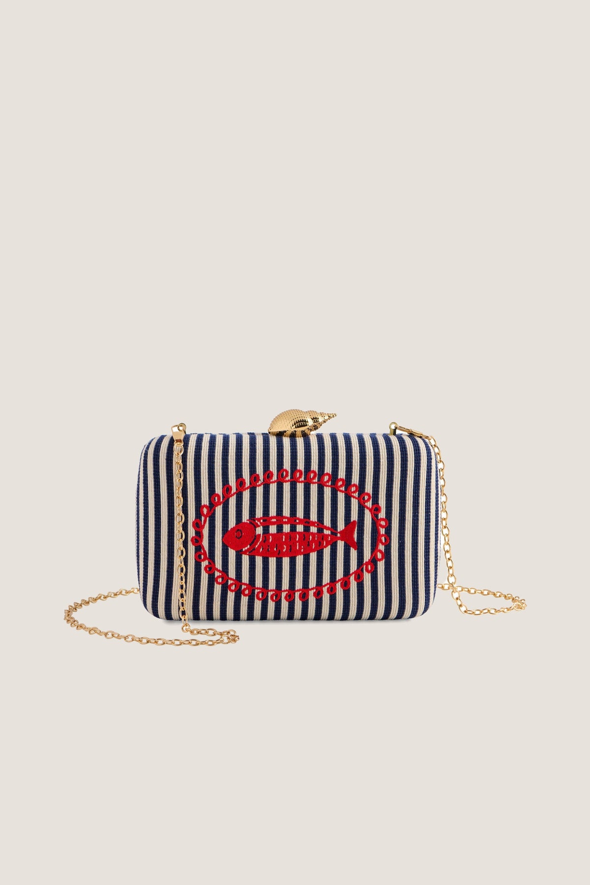 Navy Marina minaudiere clutch in navy and ivory stripe with a red fish and gold shell clasp, chain strap draped, front view on a warm greige backdrop.