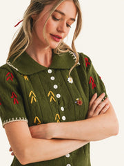 A woman wearing an olive green short-sleeve knit top with embroidered red and yellow motifs, white buttons, and a white collar, posed against a plain light background.