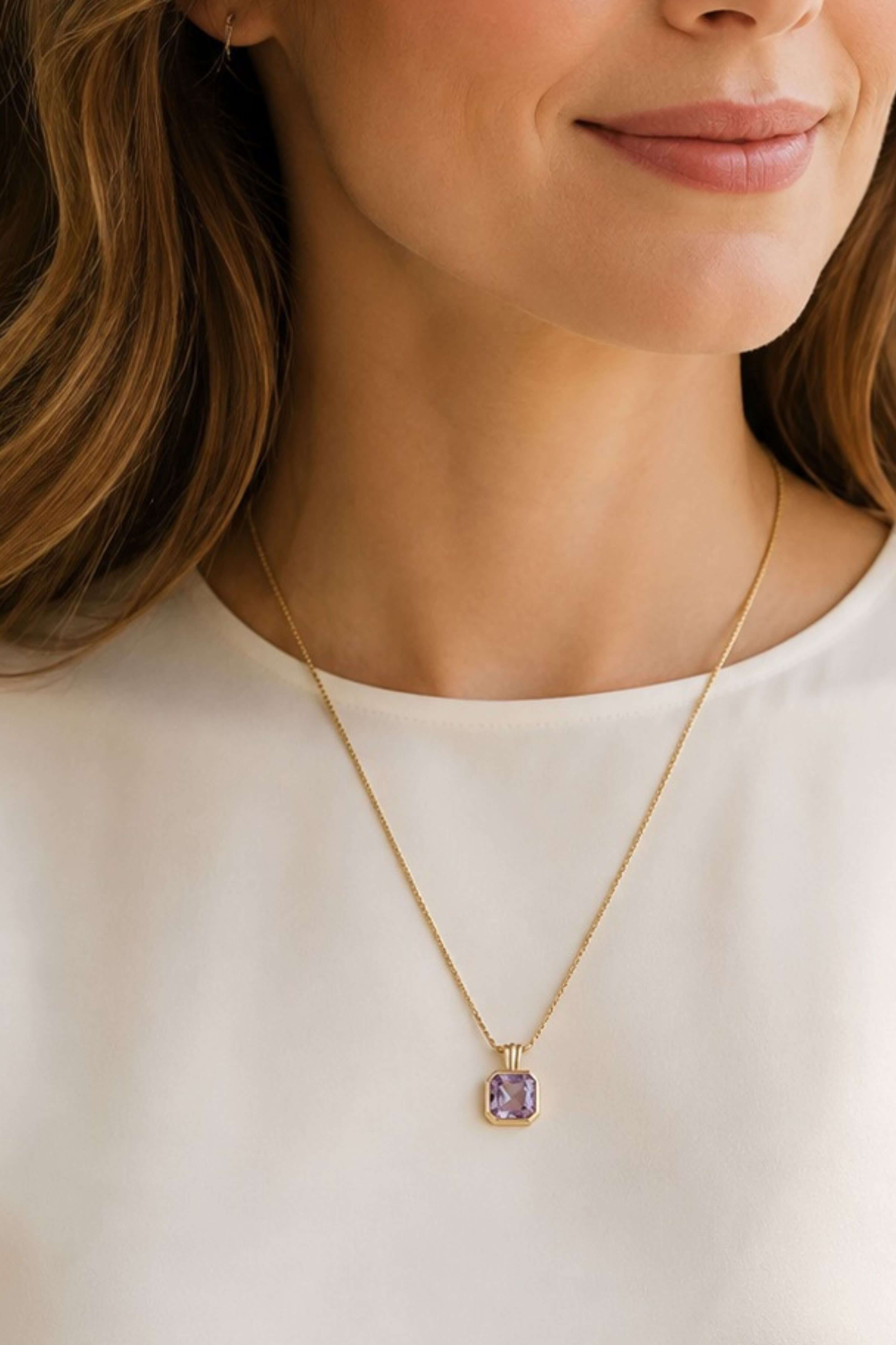 A close-up of a woman wearing a delicate gold chain necklace with a small square pendant featuring a lavender-purple gemstone, paired with a white top.