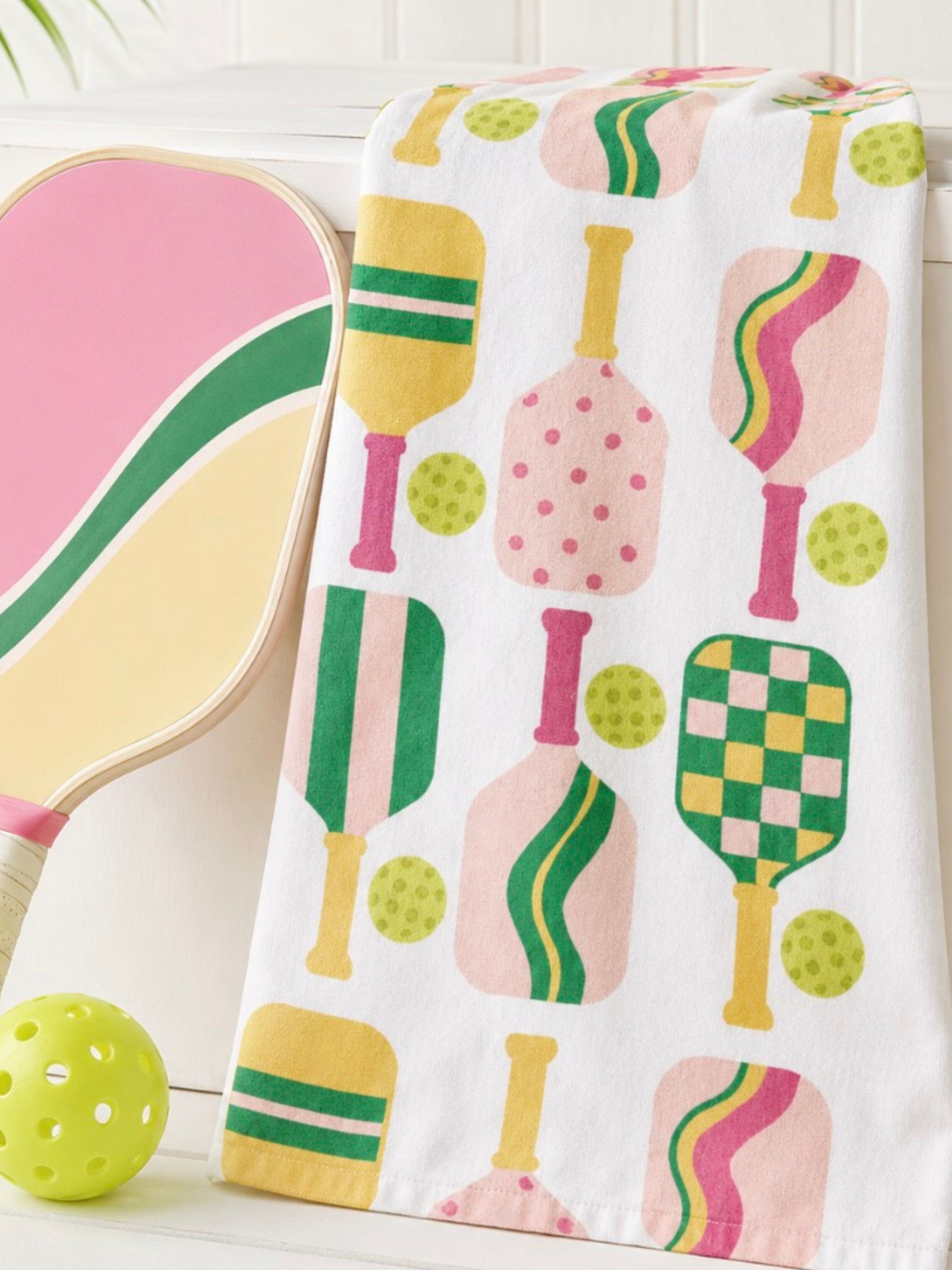A white towel with a colorful retro pattern of pickleball paddles, balls, and dots, draped next to a matching paddle and a green ball.