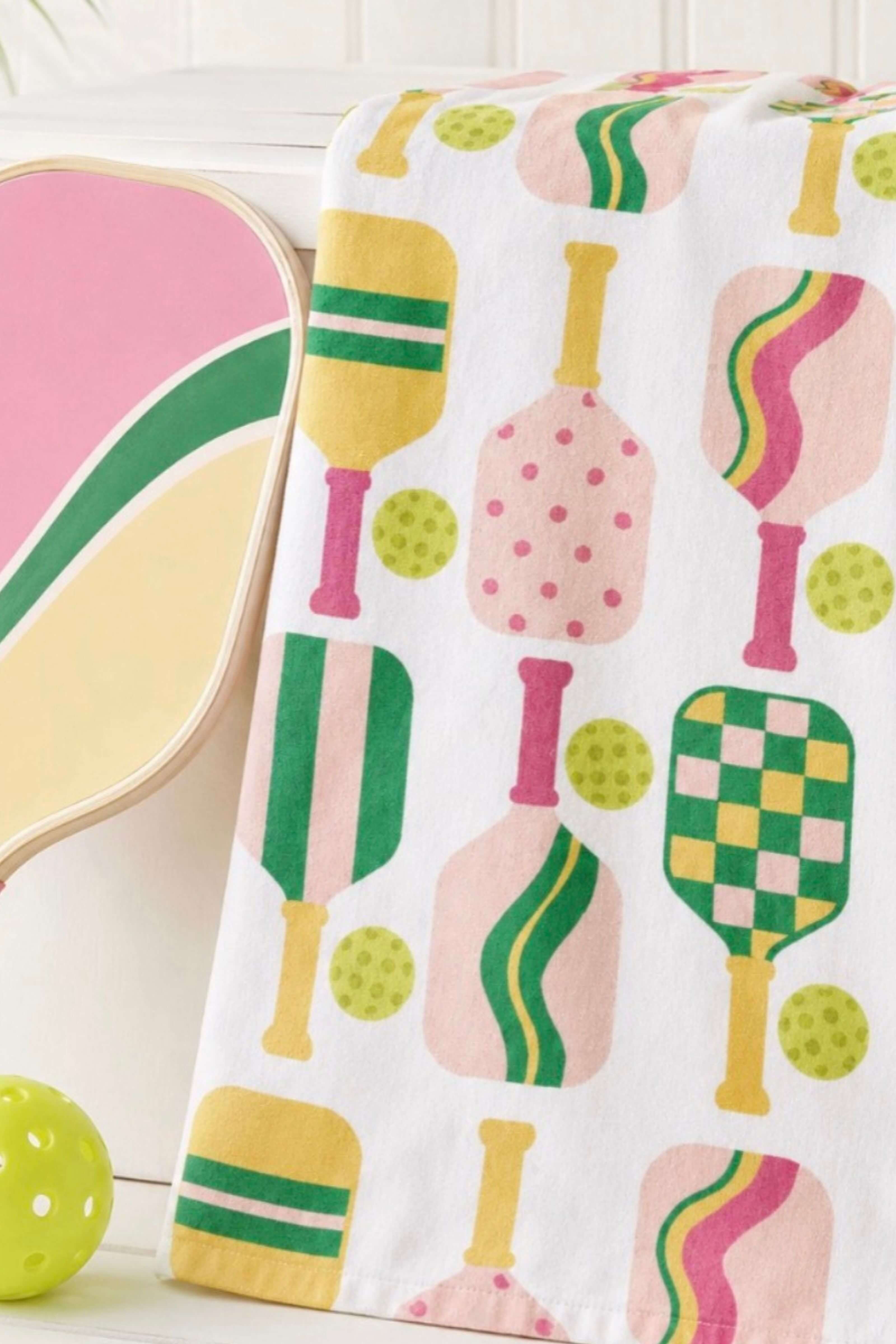 A white towel draped over a chair, featuring a colorful pattern of stylized paddle-shaped objects in pink, green, yellow, and cream. A portion of a pink and green chair is visible on the left, along with a green ball in the lower left corner.