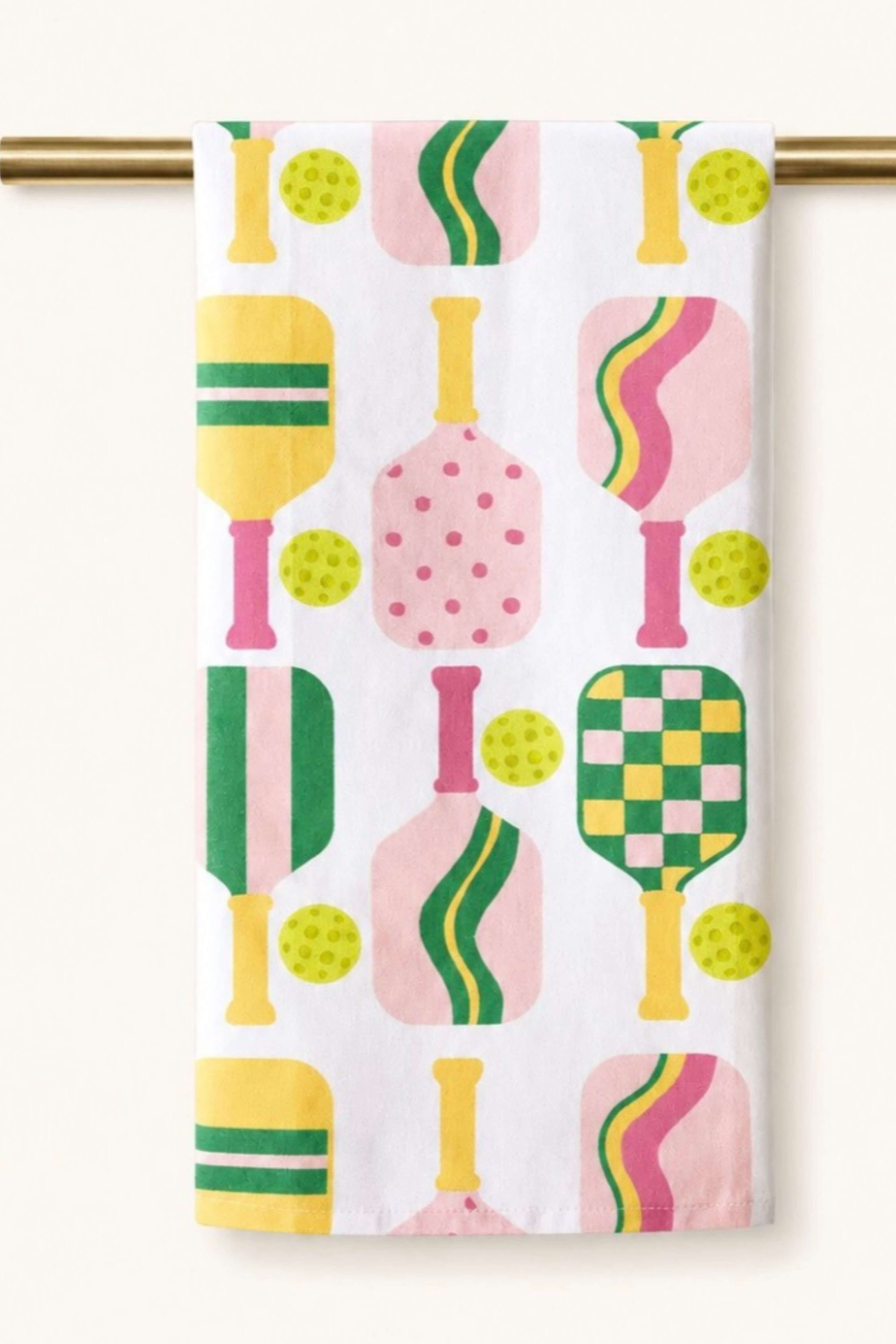 A white kitchen towel featuring a colorful retro pattern of stylized pickleball paddles and yellow-green balls, hanging from a gold-colored rod against a light beige wall.