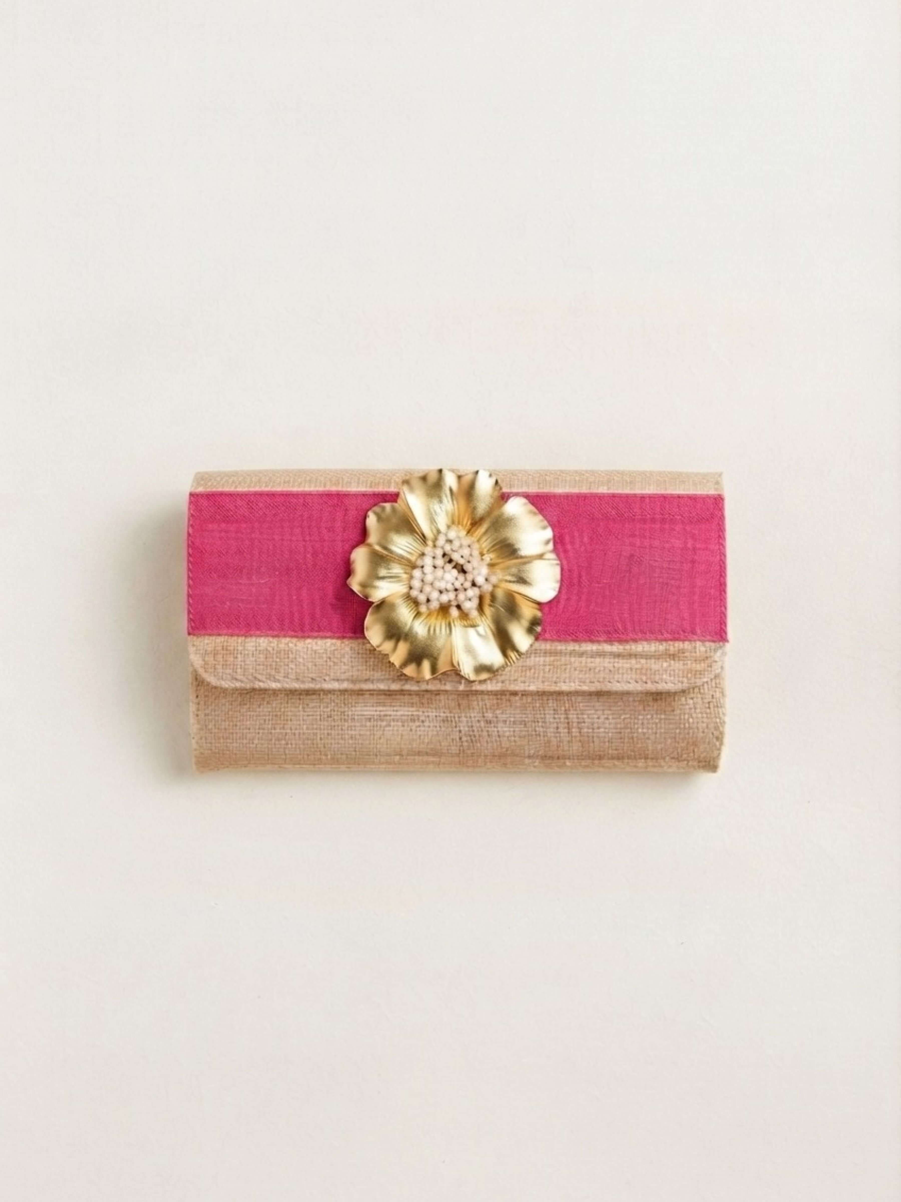 A rectangular clutch bag featuring a vibrant pink satin band across the front, a beige textured fabric base, and a large metallic gold flower embellishment at the center, set against a plain off-white background.