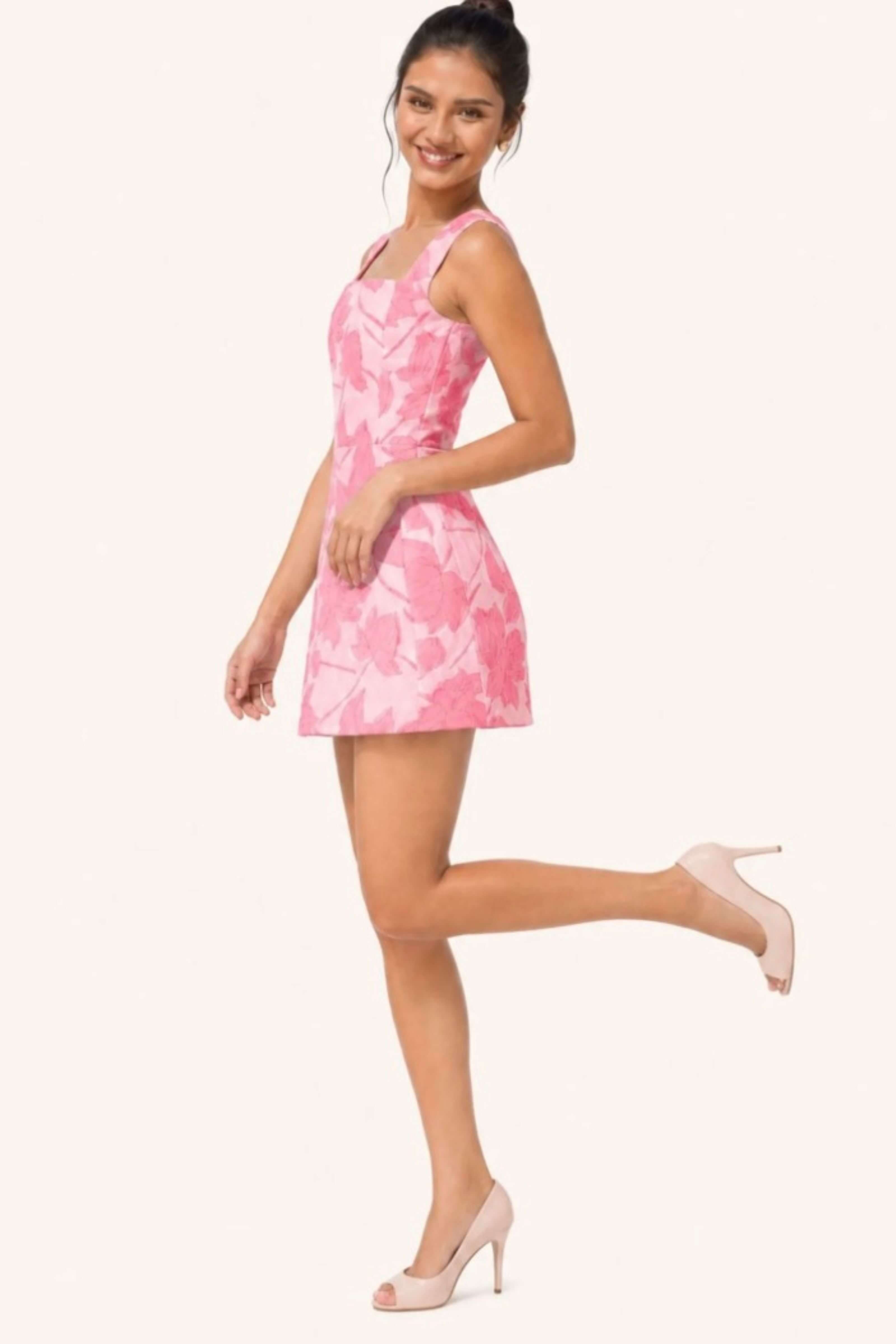 A woman modeling a sleeveless pink and white abstract patterned mini dress with beige high heels, posed against a solid off-white background.