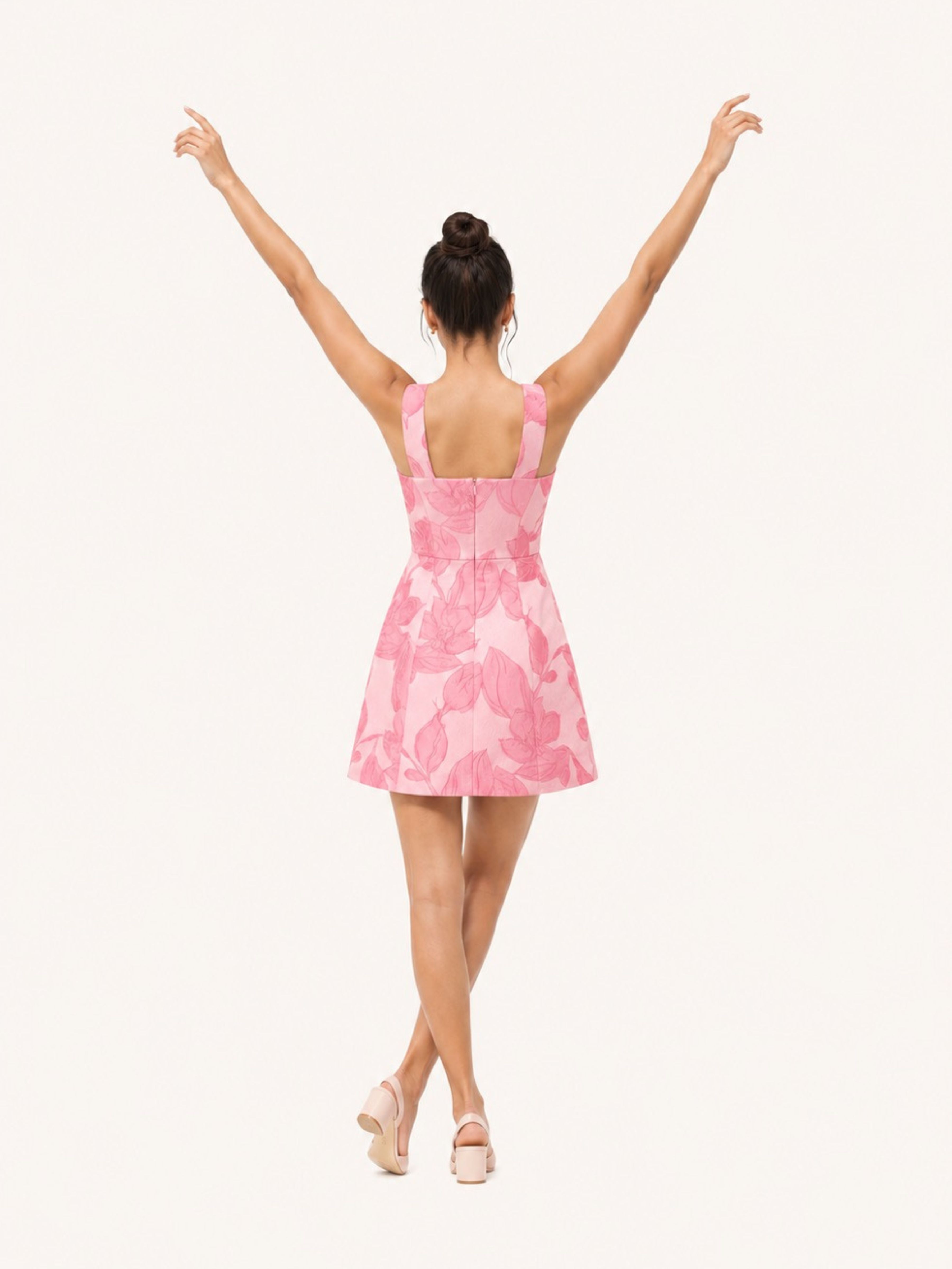 A woman viewed from behind wearing a pink sleeveless mini dress with a tonal floral pattern, standing against a plain off-white background with her arms raised.