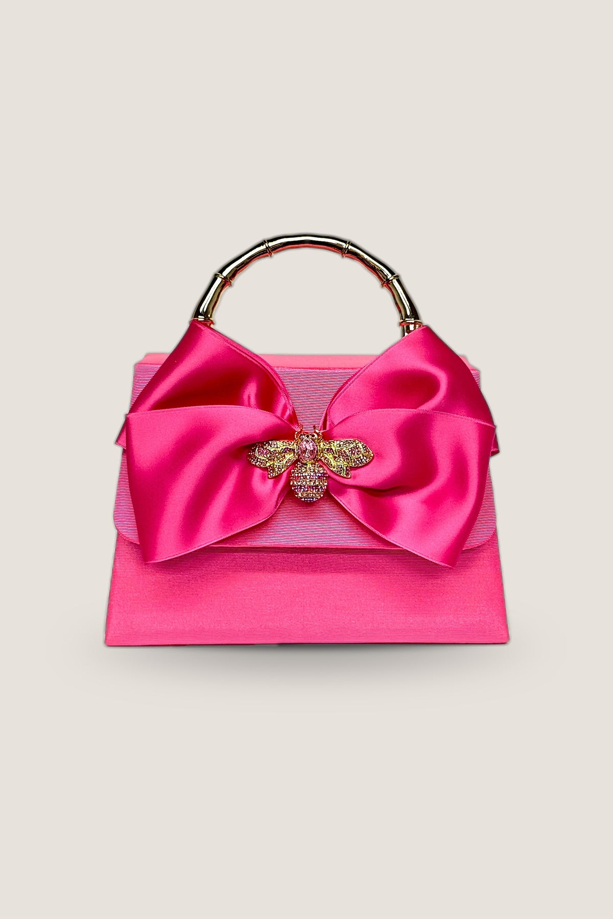 Pink Lulu handbag in hot pink with a satin bow, rhinestone bee charm and gold bamboo-style handle, front view on a warm greige backdrop.