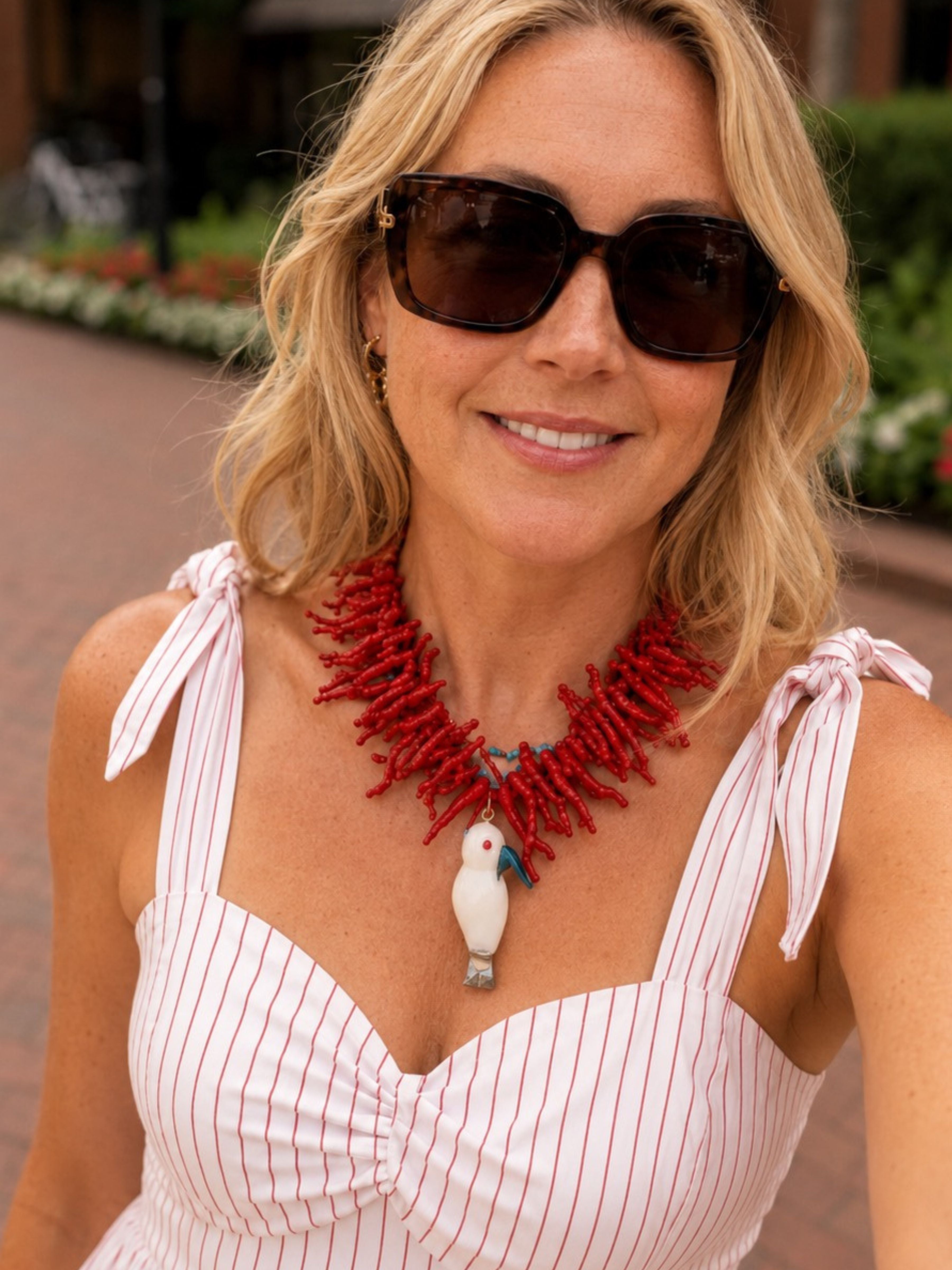 A woman wearing a chunky red coral-style necklace with a white bird pendant, paired with a white and pink striped top and dark sunglasses, photographed outdoors.