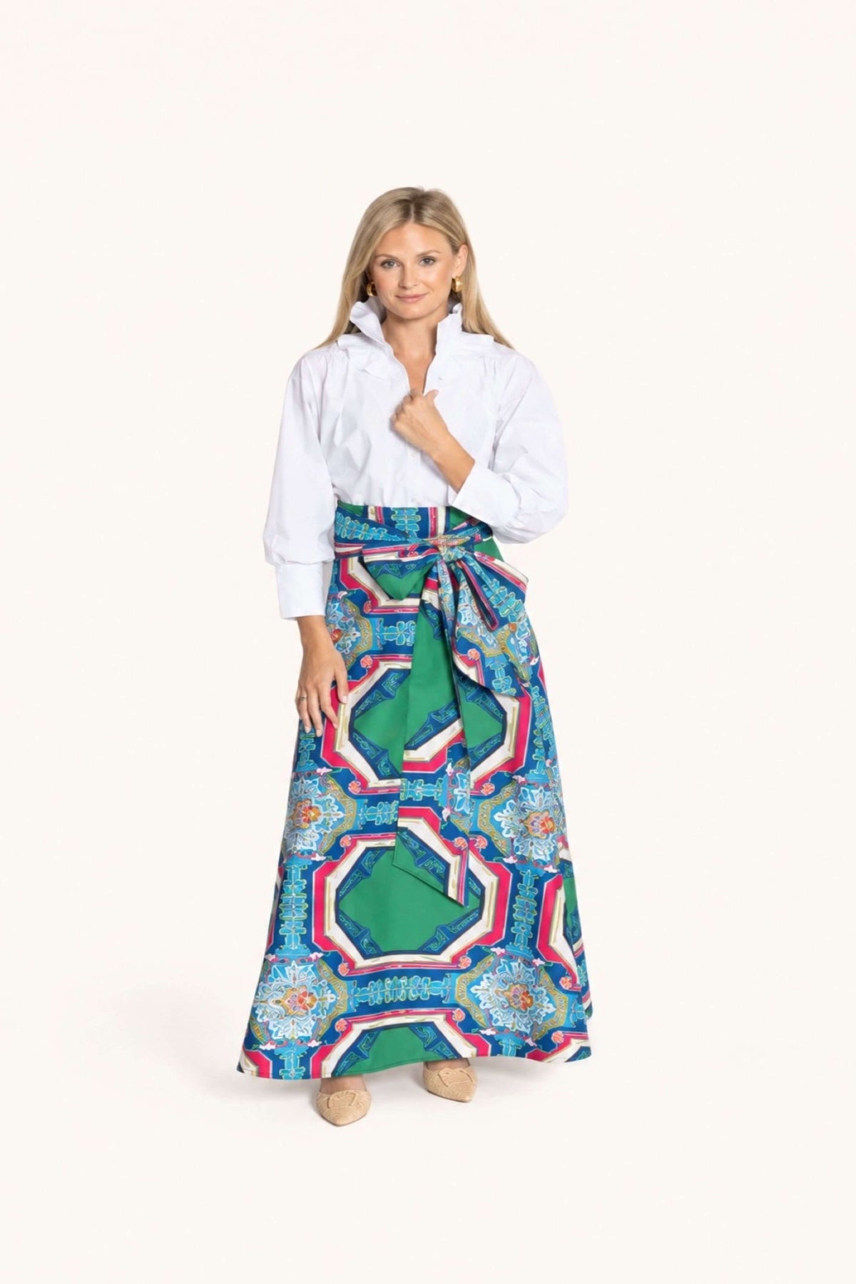 A woman modeling a long, high-waisted maxi skirt with a vibrant geometric pattern in green, blue, pink, and white, styled with a white button-up shirt against a solid off-white background.