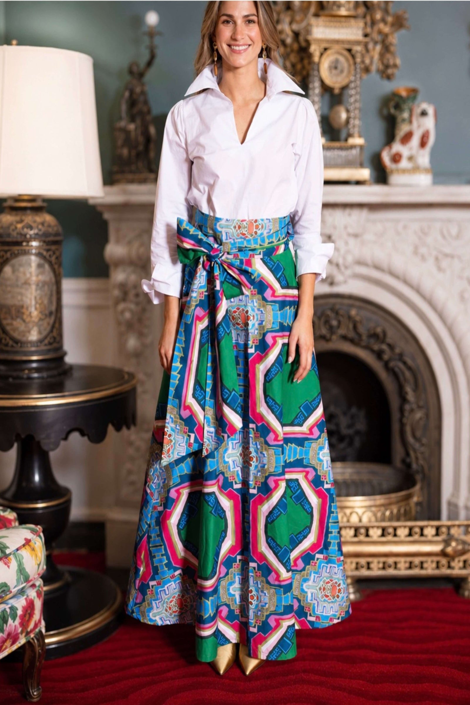 A woman stands in a decorated room wearing a white button-up shirt and a long, wide skirt with a bold geometric pattern in green, blue, pink, and white. The skirt has a matching fabric tie belt at the waist.