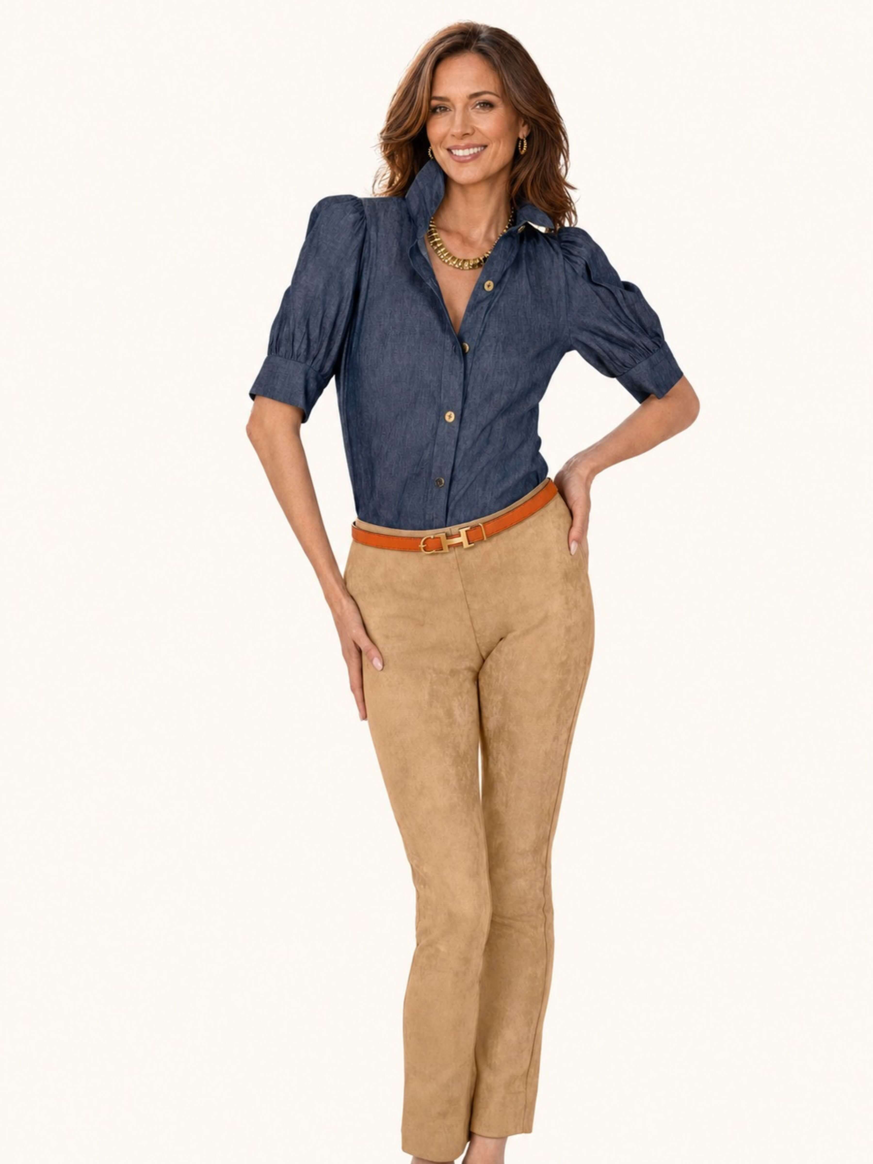 A woman modeling a dark blue chambray button-down shirt with puffed short sleeves and tan suede-like cropped pants, styled with a brown belt and gold necklace against a plain white background.