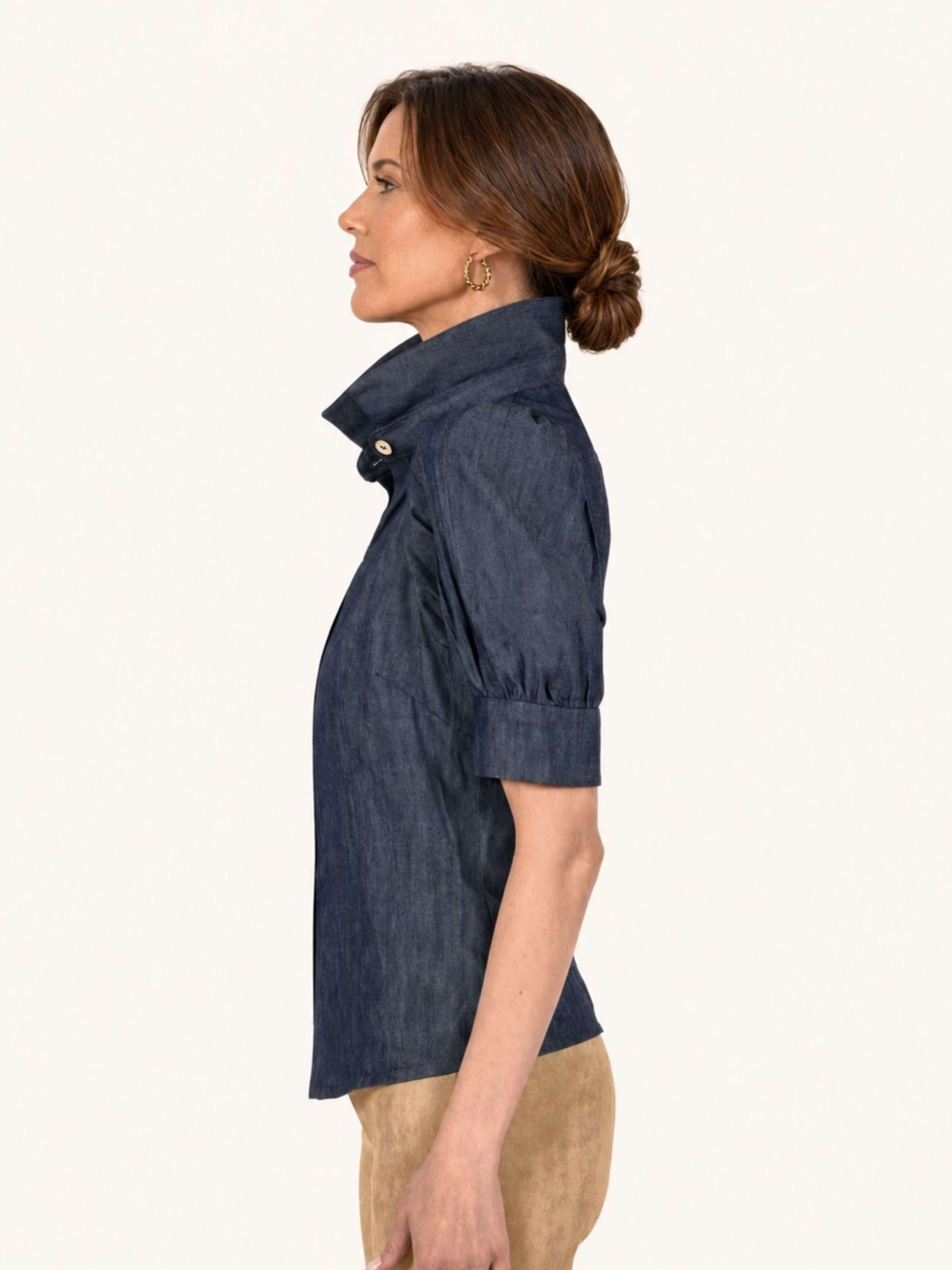 A woman shown in side profile wearing a dark indigo chambray short-sleeve shirt with a high collar and tan pants, against a solid off-white background.