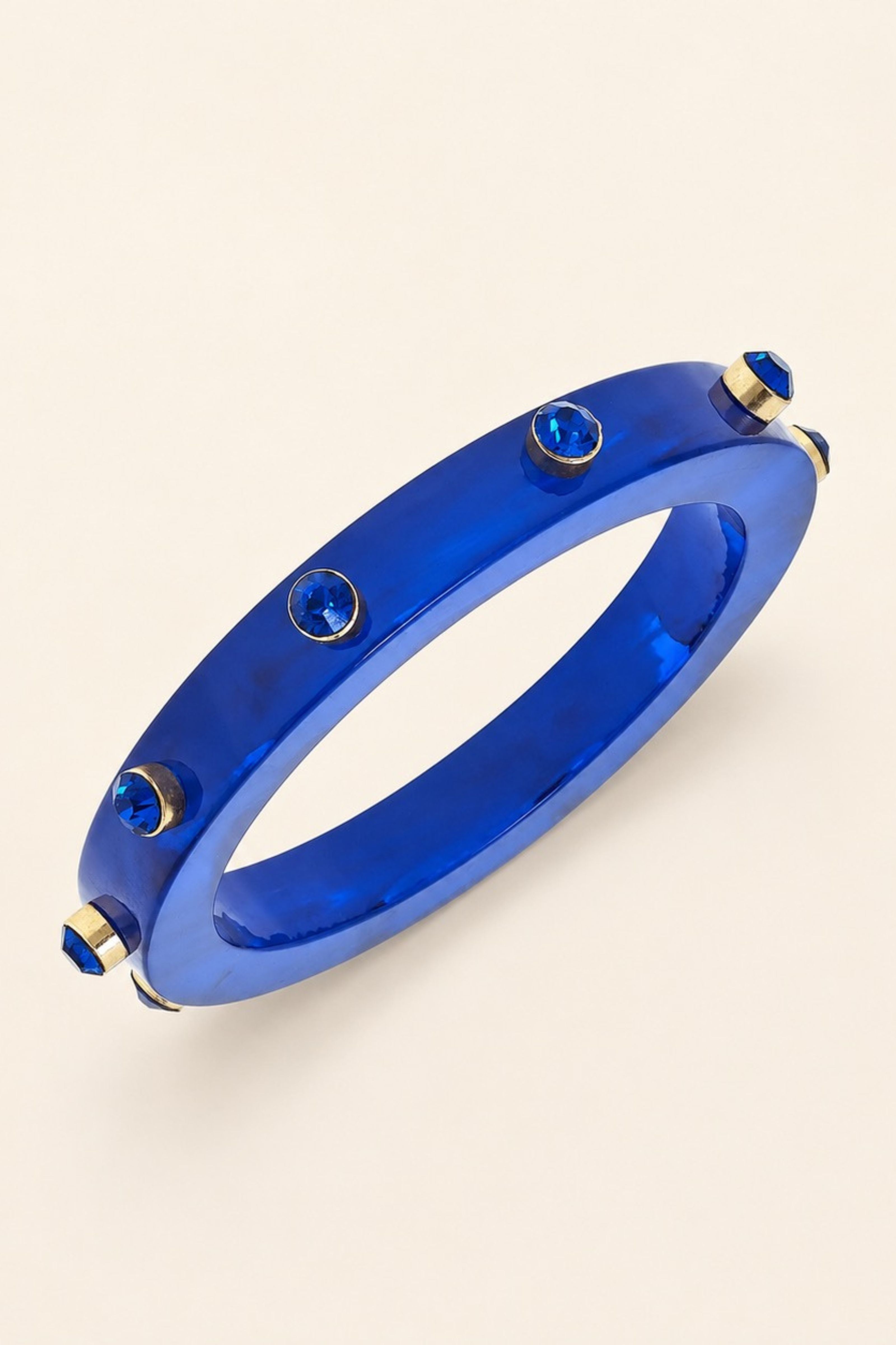 A glossy cobalt blue bangle bracelet with gold-toned hardware and small blue gemstone accents, shown on a light beige background.