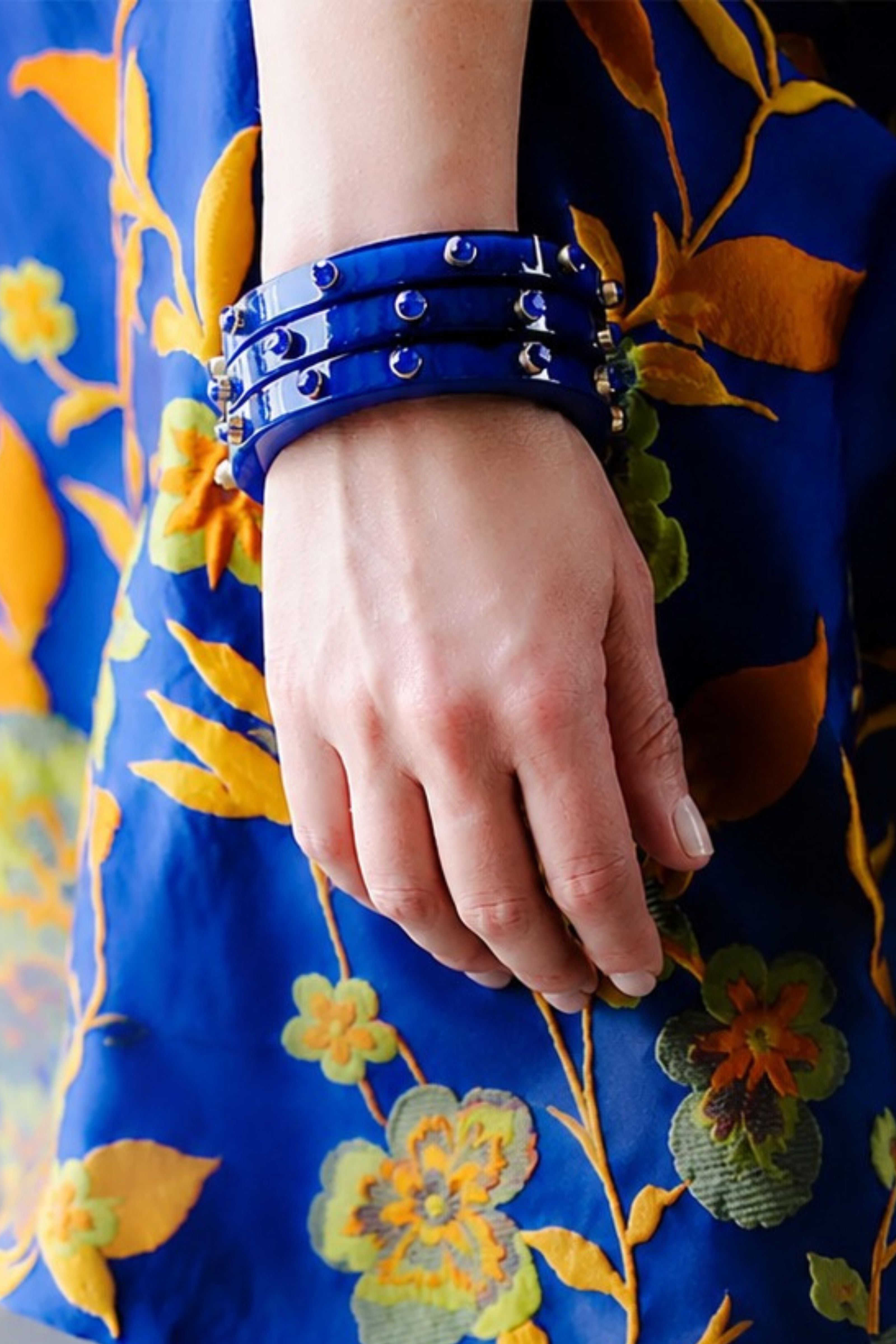 A close-up of a person's hand wearing a stack of royal blue leather bracelets with silver studs, set against a vibrant blue floral embroidered dress.