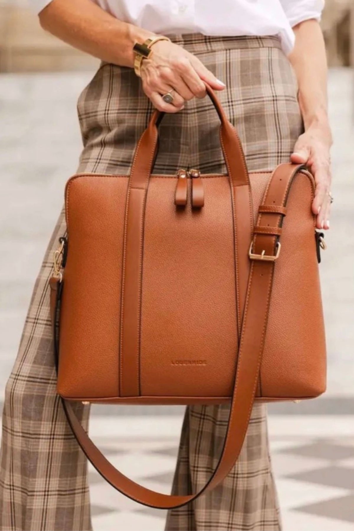 The tan Rhodes Laptop Bag held by its top handles against plaid trousers.