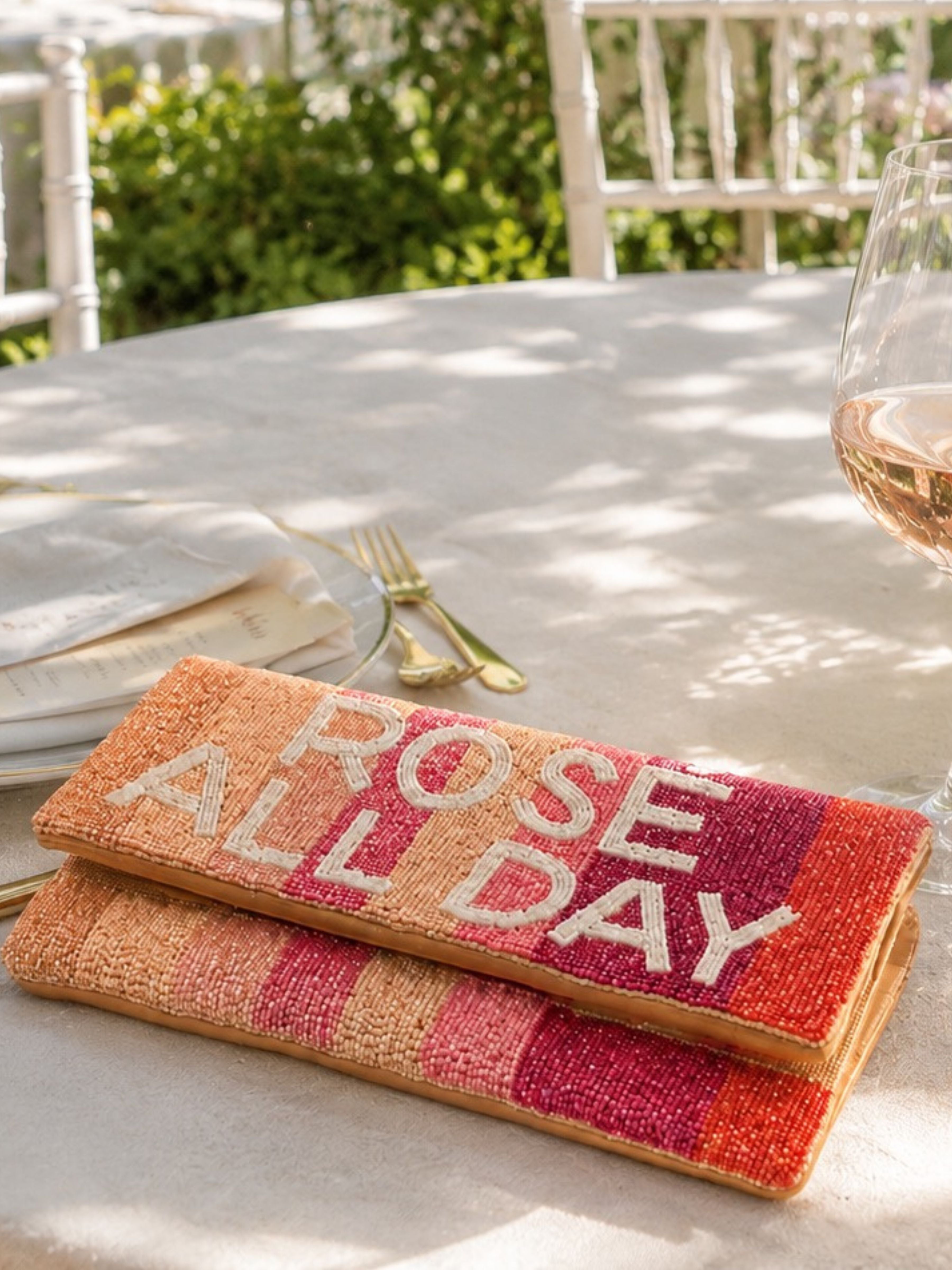A woven rectangular placemat or napkin with the phrase “ROSE ALL DAY” in large white letters, layered on top of a matching striped textile, set on an outdoor dining table with a wine glass, folded napkin, and gold cutlery nearby.