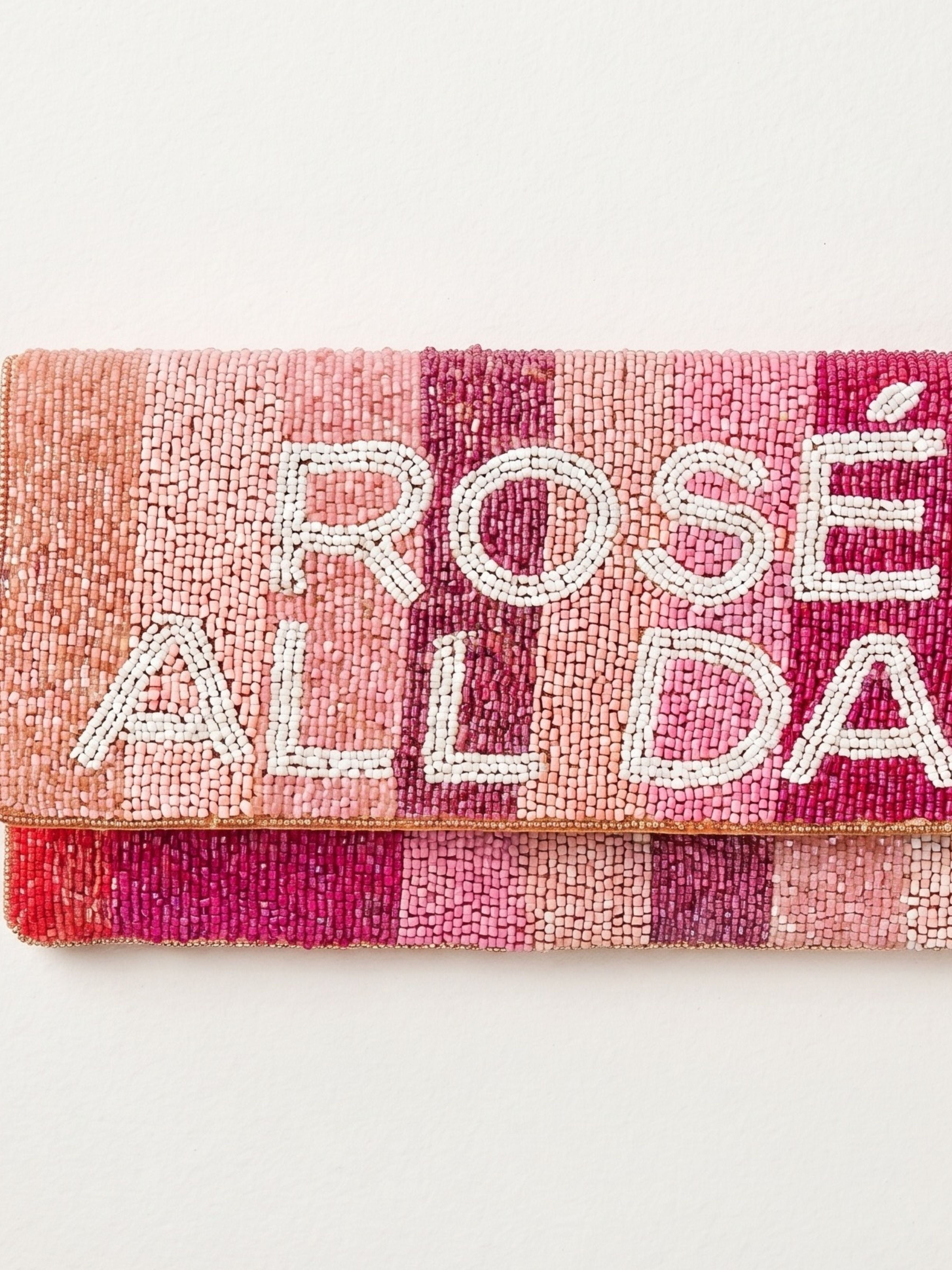 A close-up of a beaded clutch bag with a folded flap, featuring the text 'ROSÉ ALL DAY' in white bead lettering over a striped pattern of pink, coral, and magenta tones, on a plain off-white background.