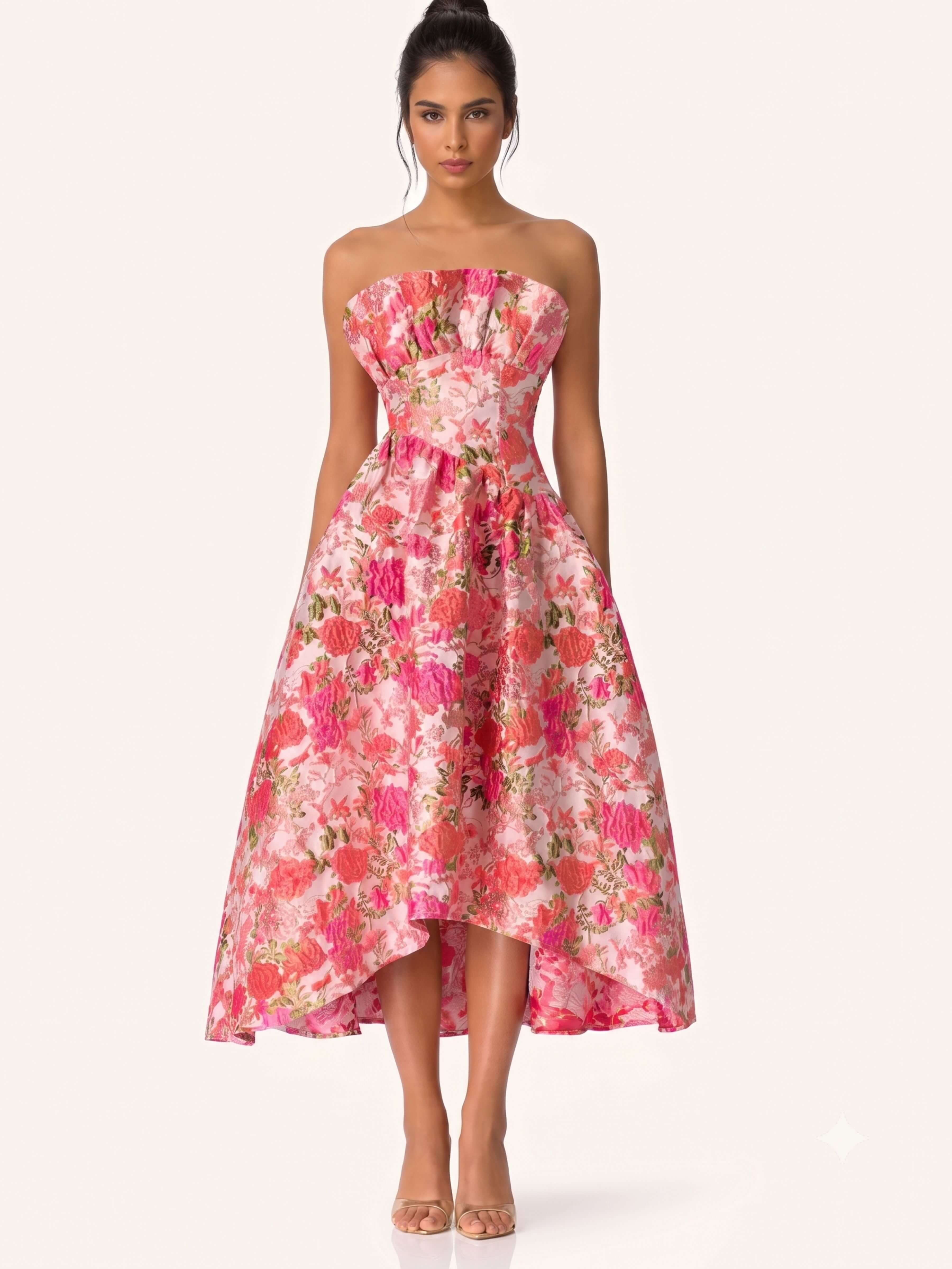 A woman modeling a strapless high-low midi dress with a pink and red floral print, set against a solid pale pink background.