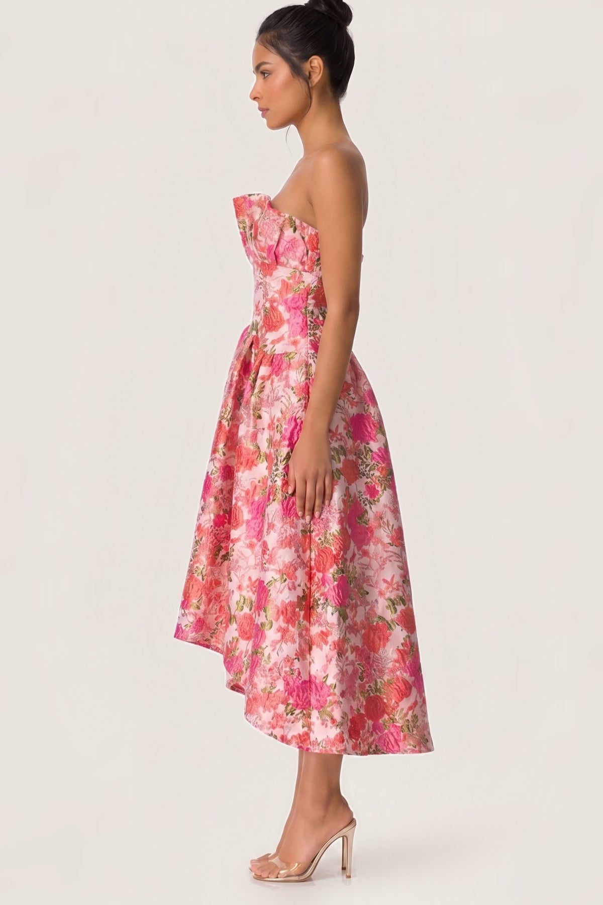 A woman modeling a strapless pink floral midi dress with a fitted bodice and flared skirt, shown in side profile against a plain light background.