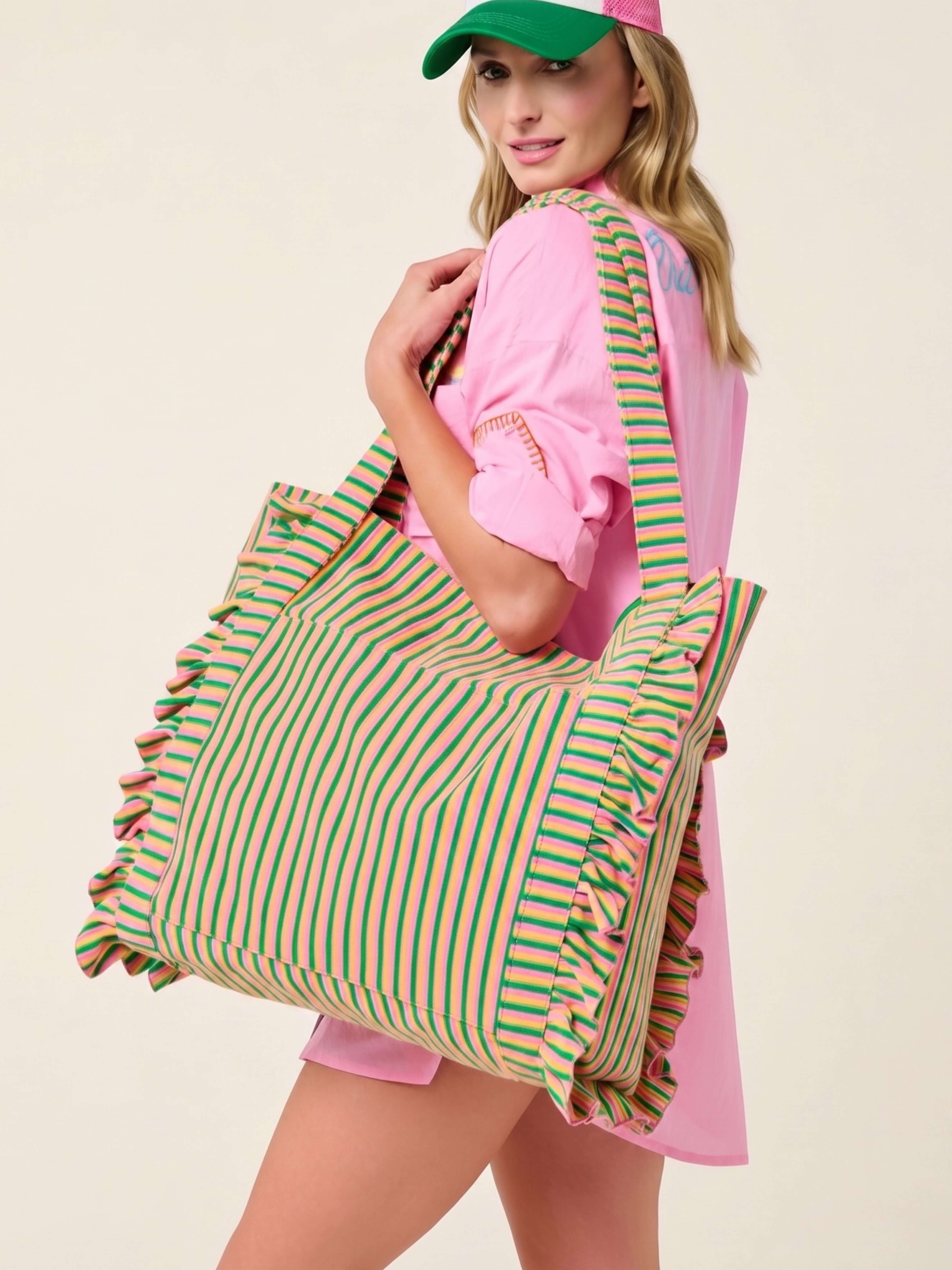 A woman wearing a pink dress and green cap carries a large striped tote bag with green, pink, and yellow vertical stripes and ruffled edges against a plain light beige background.