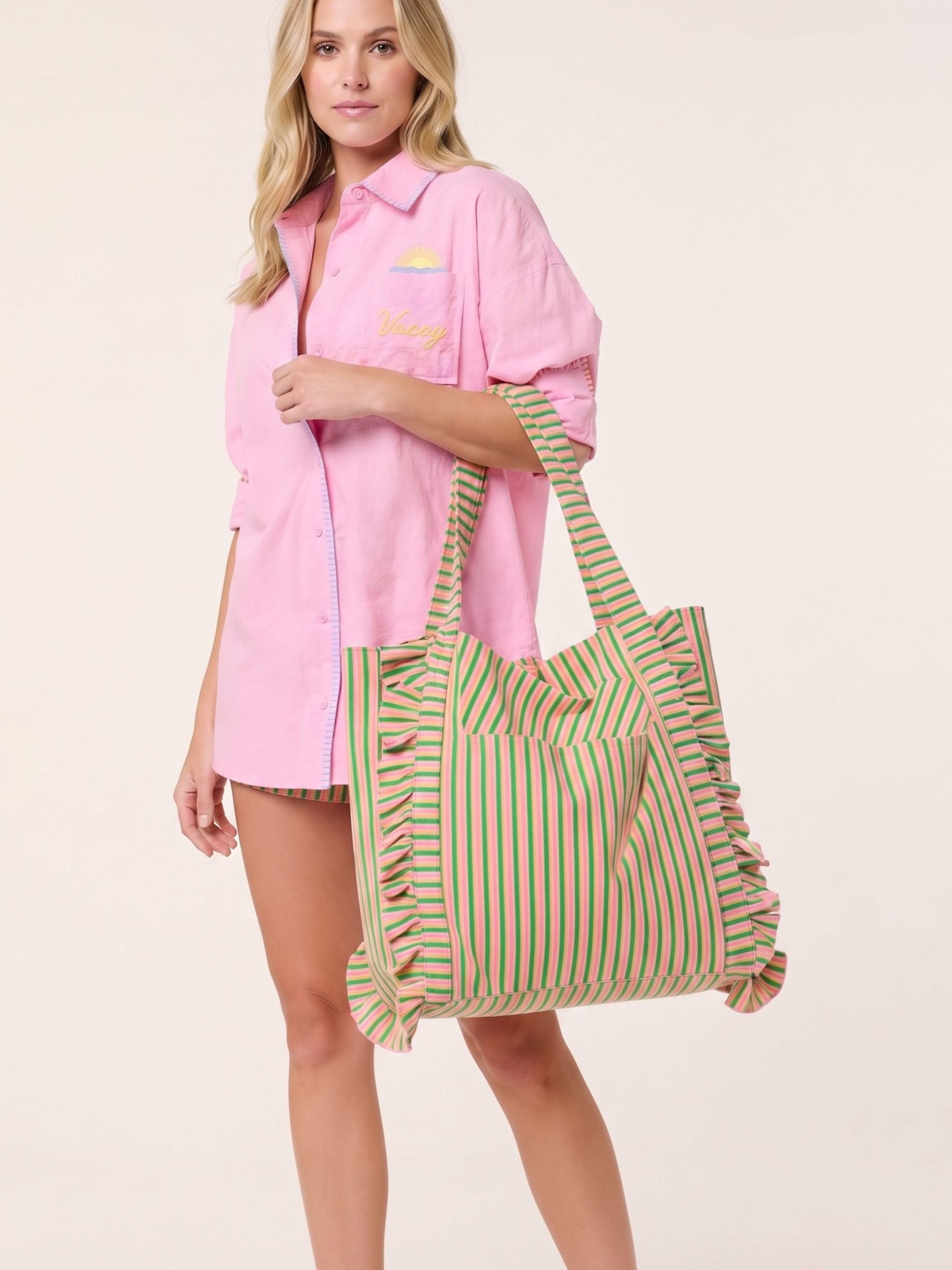 A woman models a pink button-up shirt dress and carries a large striped tote bag with green, yellow, and pink vertical stripes and ruffle trim, against a plain light beige background.