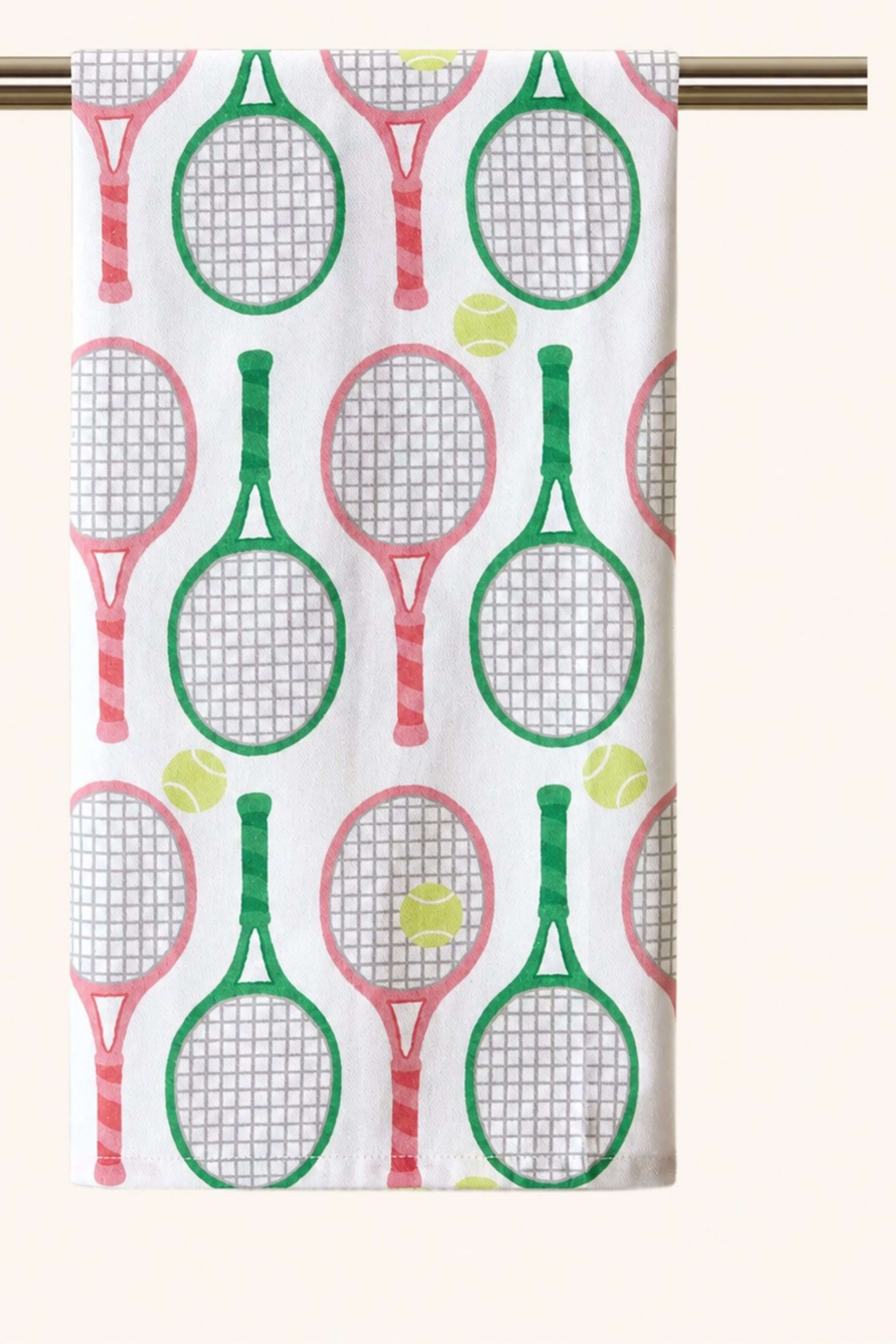A white kitchen towel featuring a repeating pattern of green and pink tennis rackets and yellow tennis balls, hanging from a gold-colored rod against a light beige background.
