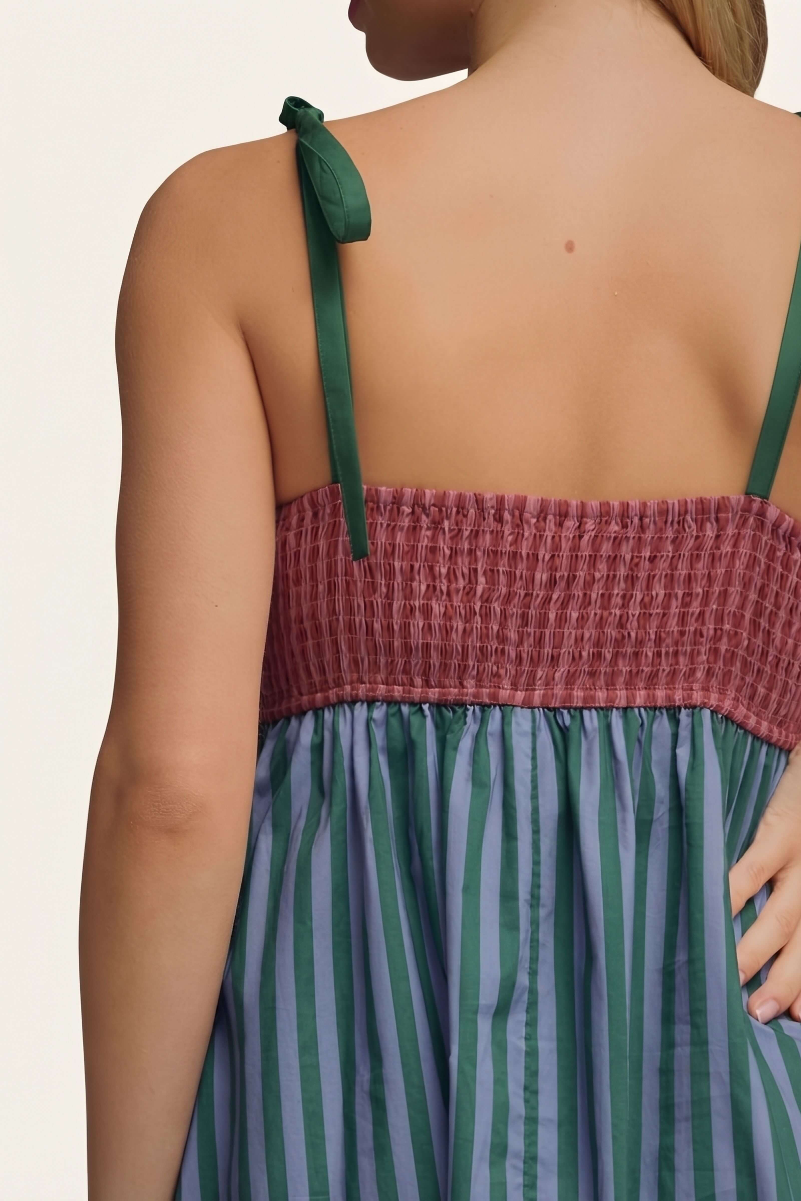 Close-up back view of a woman wearing a sleeveless dress with dark green tie straps, a smocked dusty rose band, and green and blue vertical stripes.
