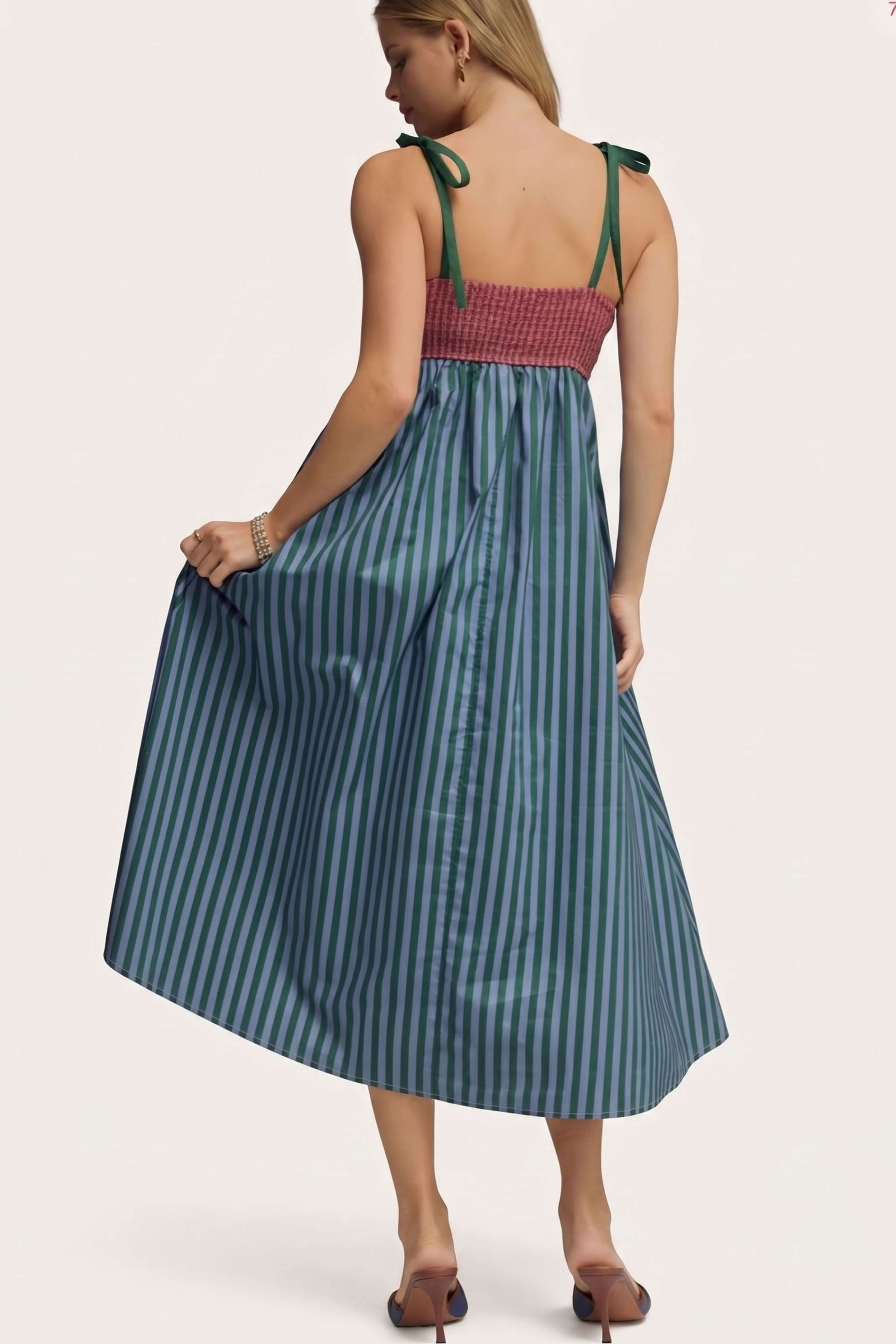 A woman modeling a sleeveless midi dress from the back, featuring a smocked dusty rose bodice with green tie straps and a blue-green and teal vertically striped skirt, set against a plain off-white background.