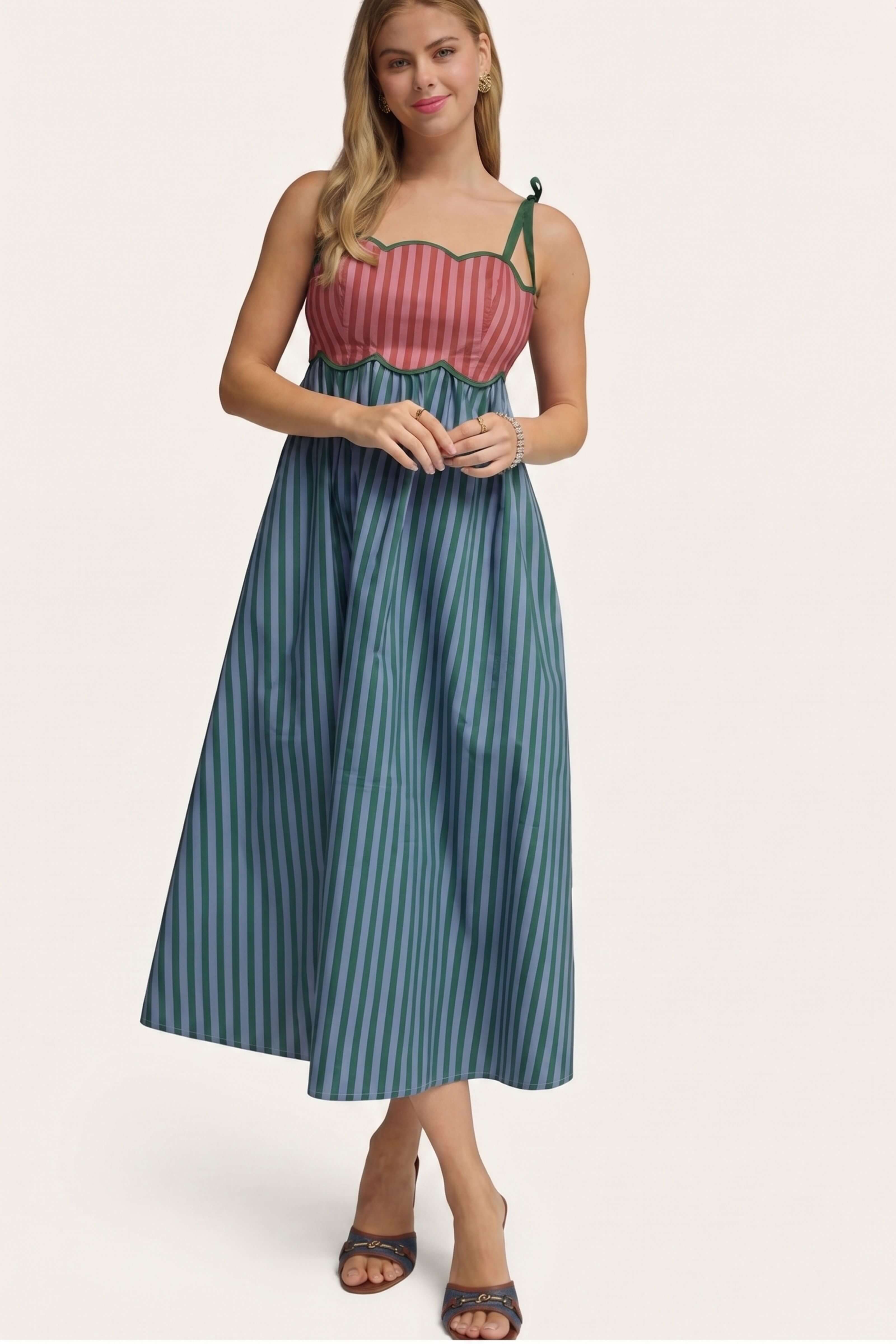 A woman modeling a sleeveless midi dress with a pink and red striped bodice and teal and blue vertical striped skirt, paired with brown heeled sandals, against a light beige studio background.