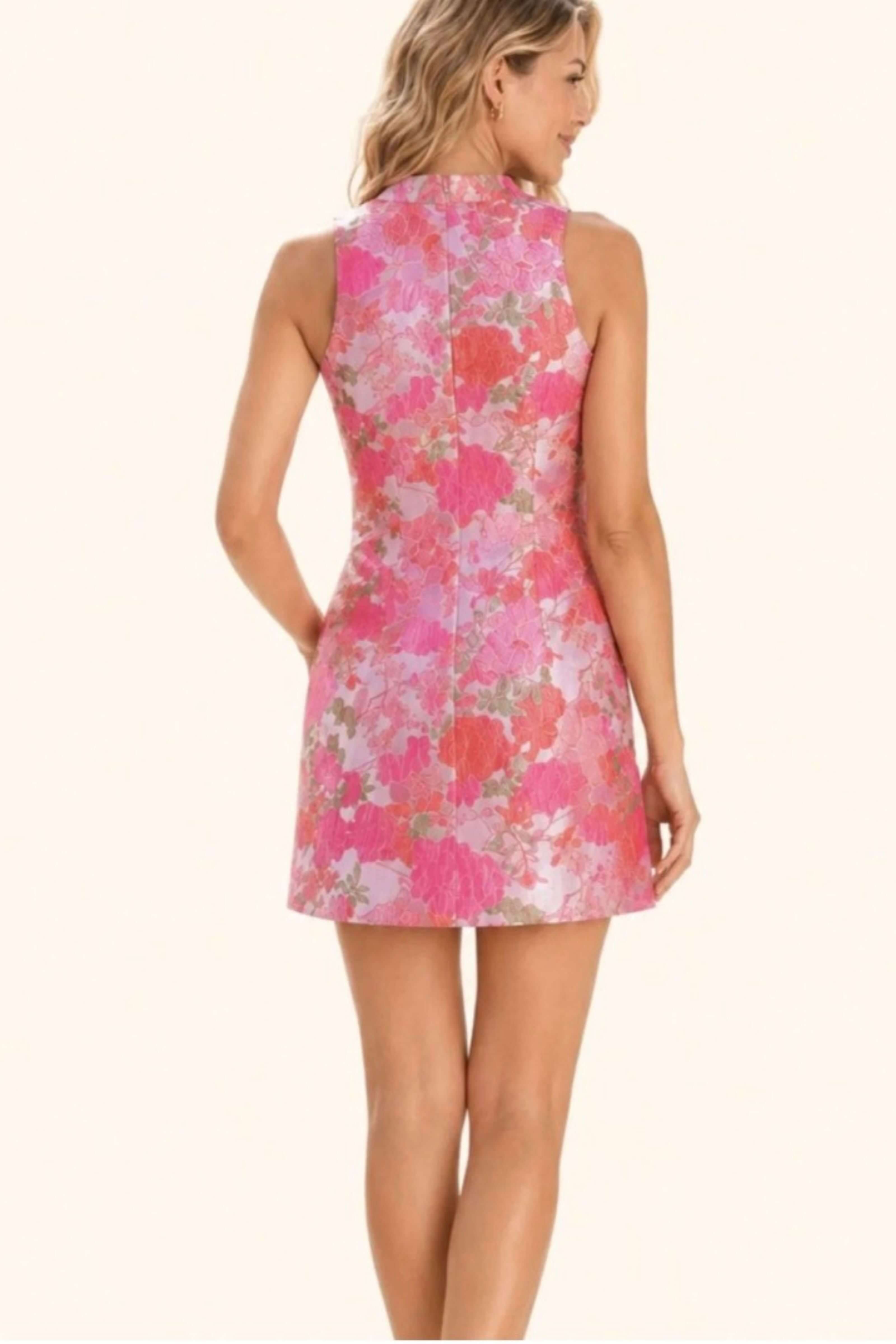 A woman shown from the back wearing a sleeveless high-neck mini dress with a pink, coral, and white floral pattern on a light background.