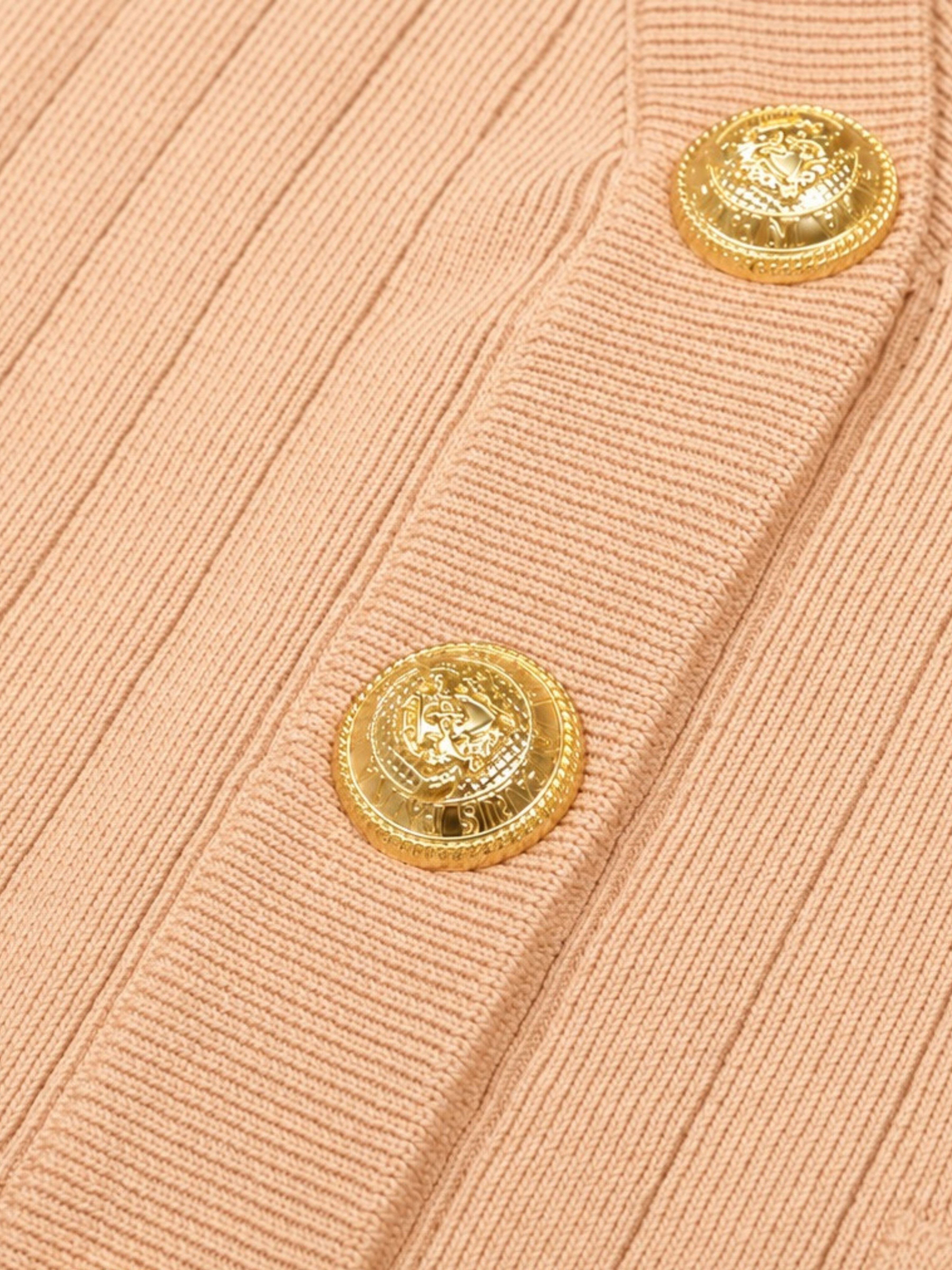 Close-up of a ribbed knit cardigan in a soft peach-pink color, featuring two prominent round gold-tone buttons with an embossed crest design.