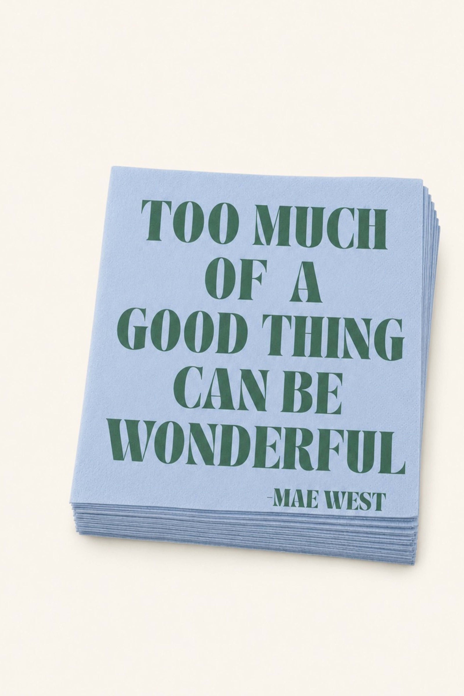 A stack of light blue paper napkins featuring the quote 'TOO MUCH OF A GOOD THING CAN BE WONDERFUL -MAE WEST' printed in dark green serif lettering, set against a solid off-white background.