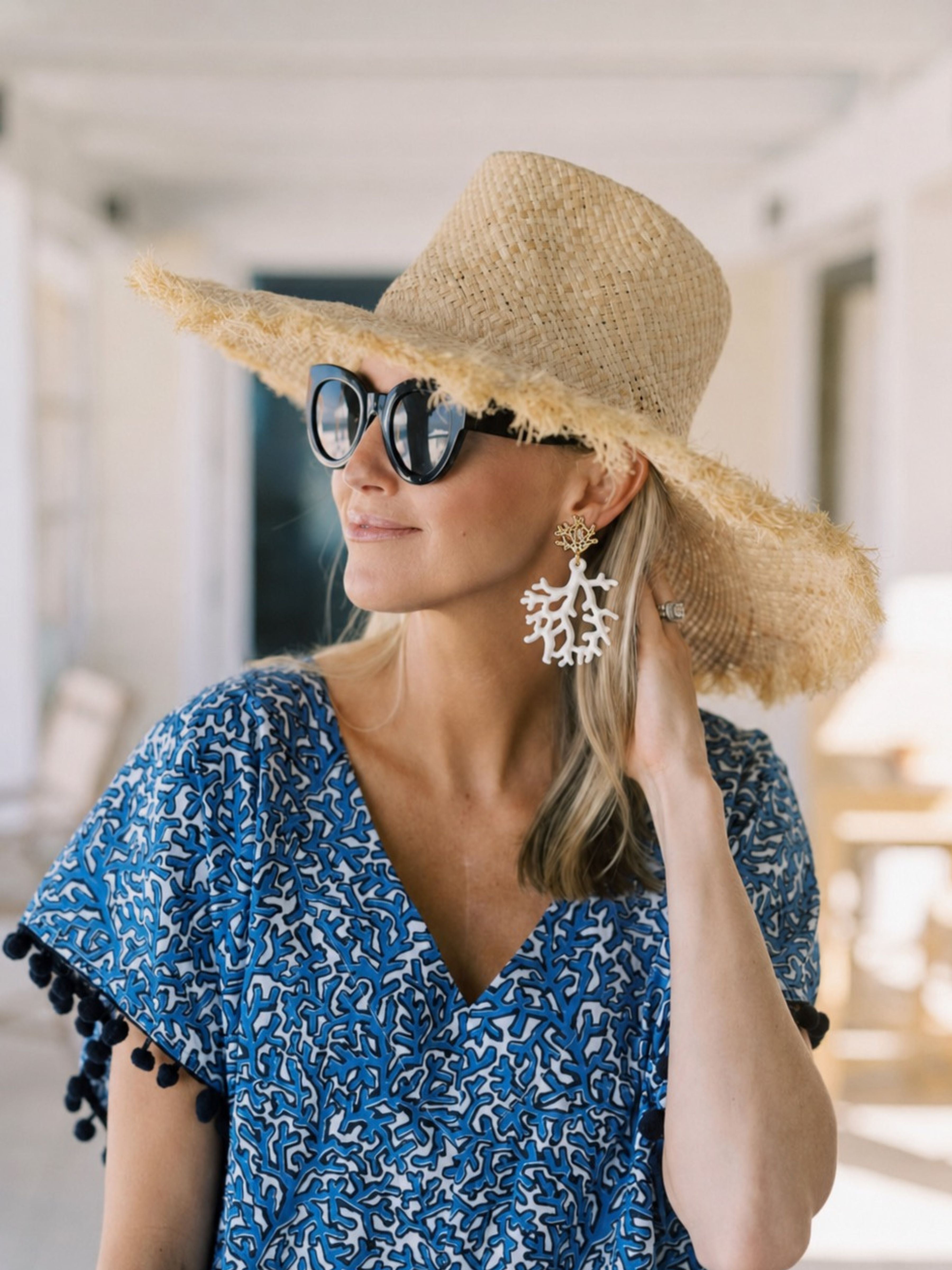 A woman wearing a wide-brimmed straw hat, black sunglasses, and a blue and white patterned top with pom-pom trim, photographed in a bright indoor setting.