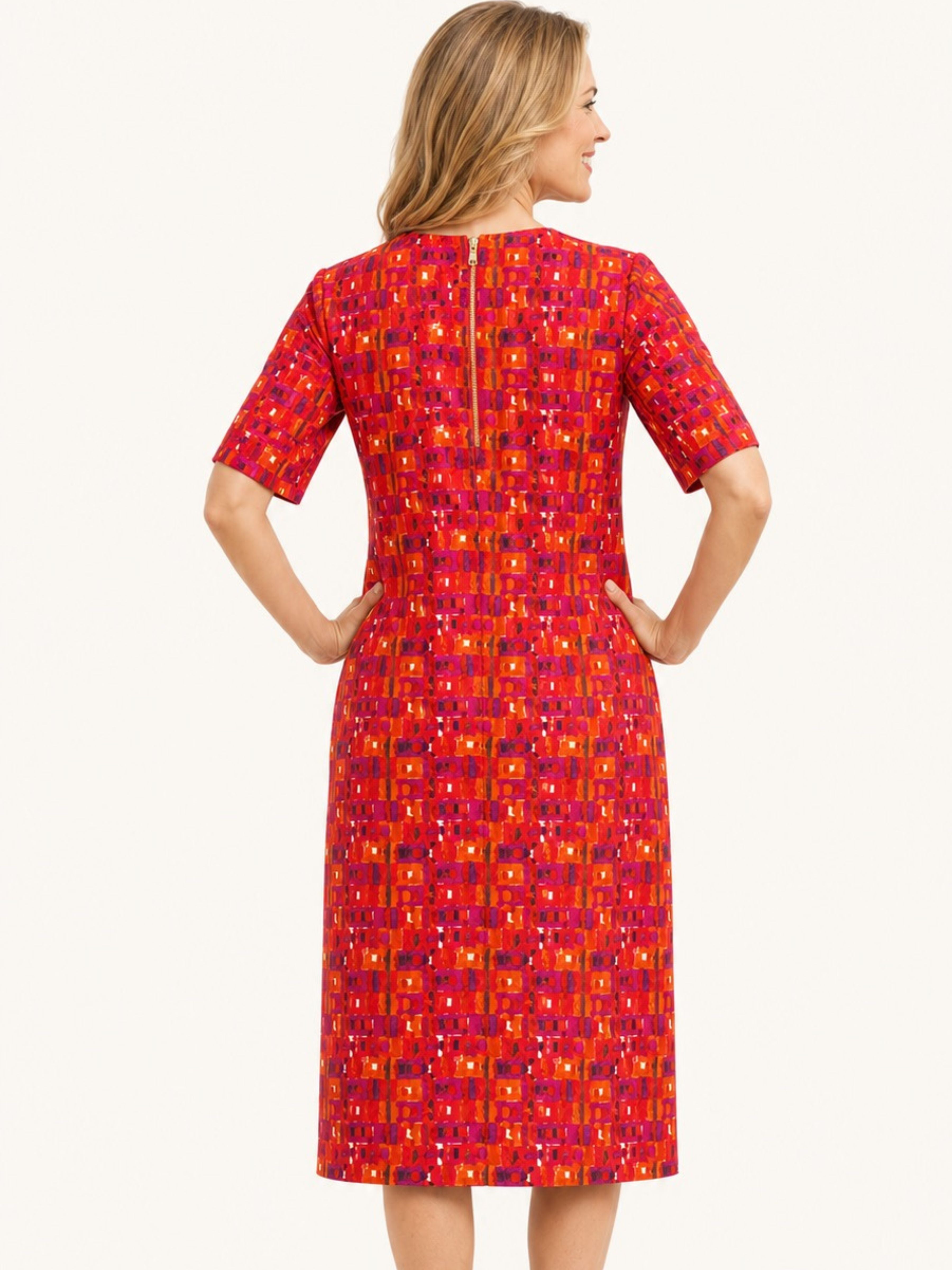 A woman shown from the back wearing a short-sleeve knee-length dress with a bold geometric pattern in red, orange, pink, and purple on a light off-white background.