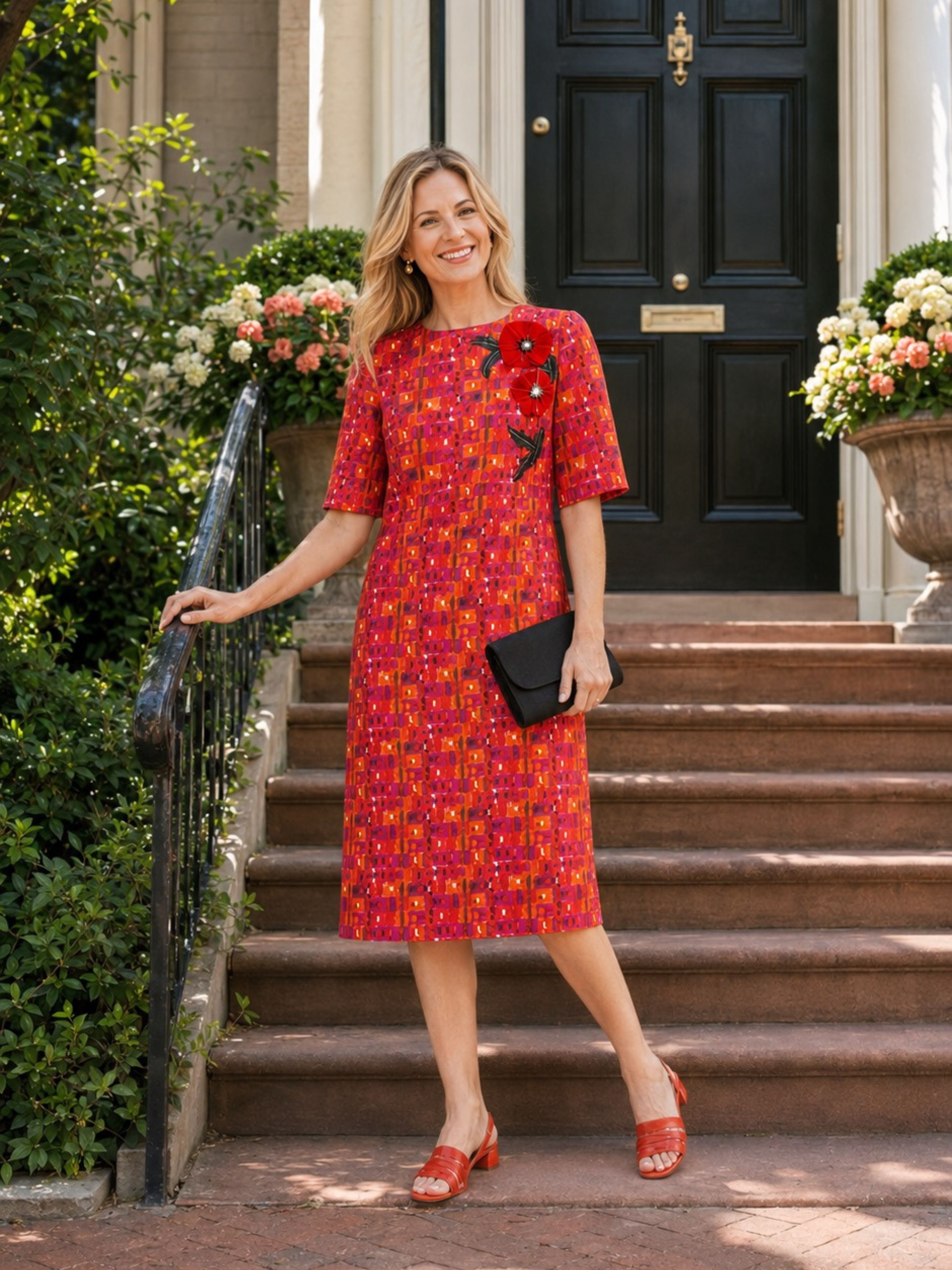 A woman stands on stone steps in front of a black front door, wearing a red knee-length dress with an orange and yellow floral pattern, short sleeves, and a black floral embellishment on the chest. She holds a small black clutch and wears red heeled sandals.