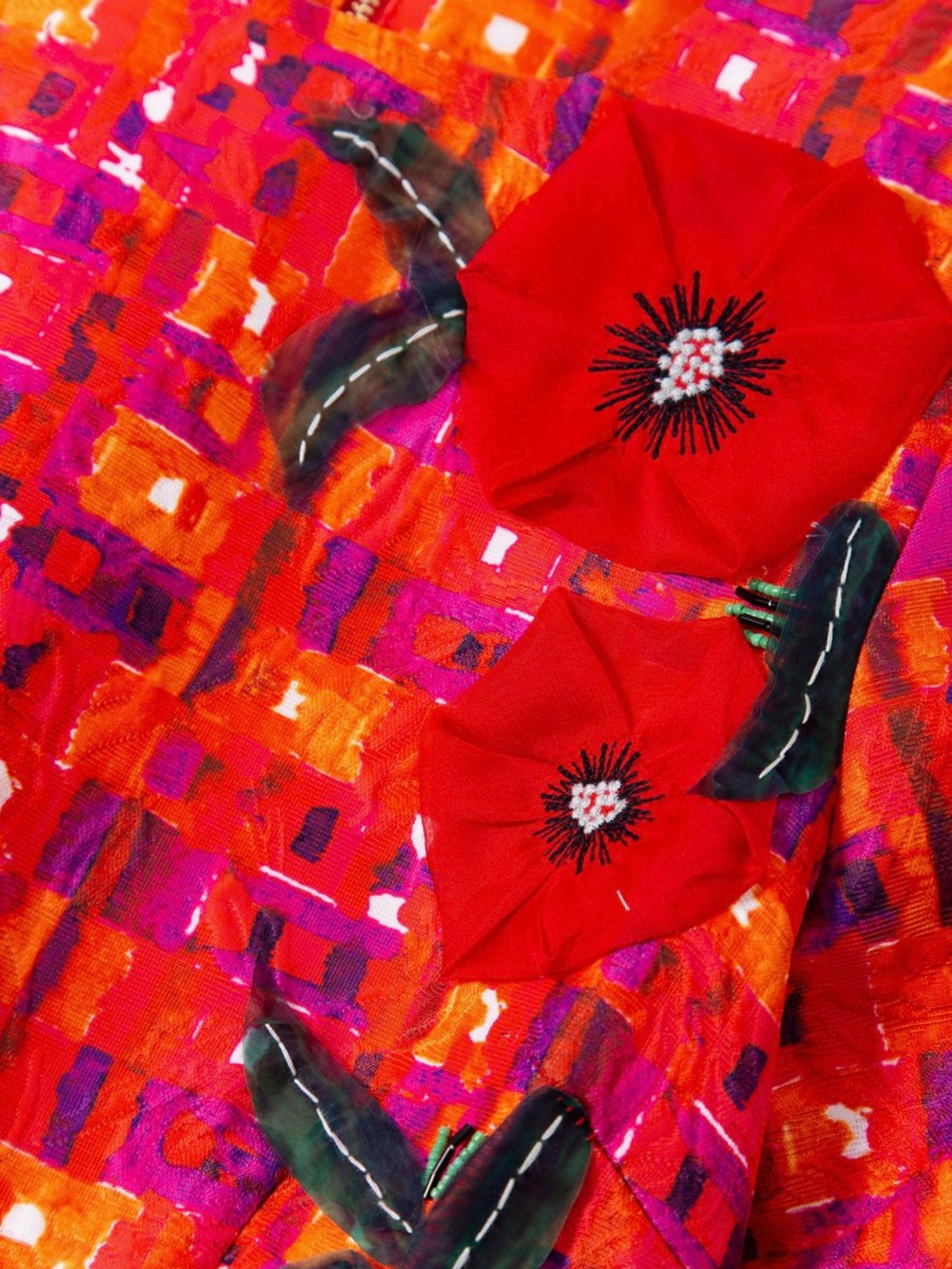 A close-up of a vibrant textile or fabric featuring two large red poppy flowers with black centers and dark green leaves, layered over a highly textured, abstract background of orange, red, pink, and purple brushstrokes.