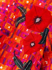 A close-up of a vibrant textile or fabric featuring two large red poppy flowers with black centers and dark green leaves, layered over a highly textured, abstract background of orange, red, pink, and purple brushstrokes.