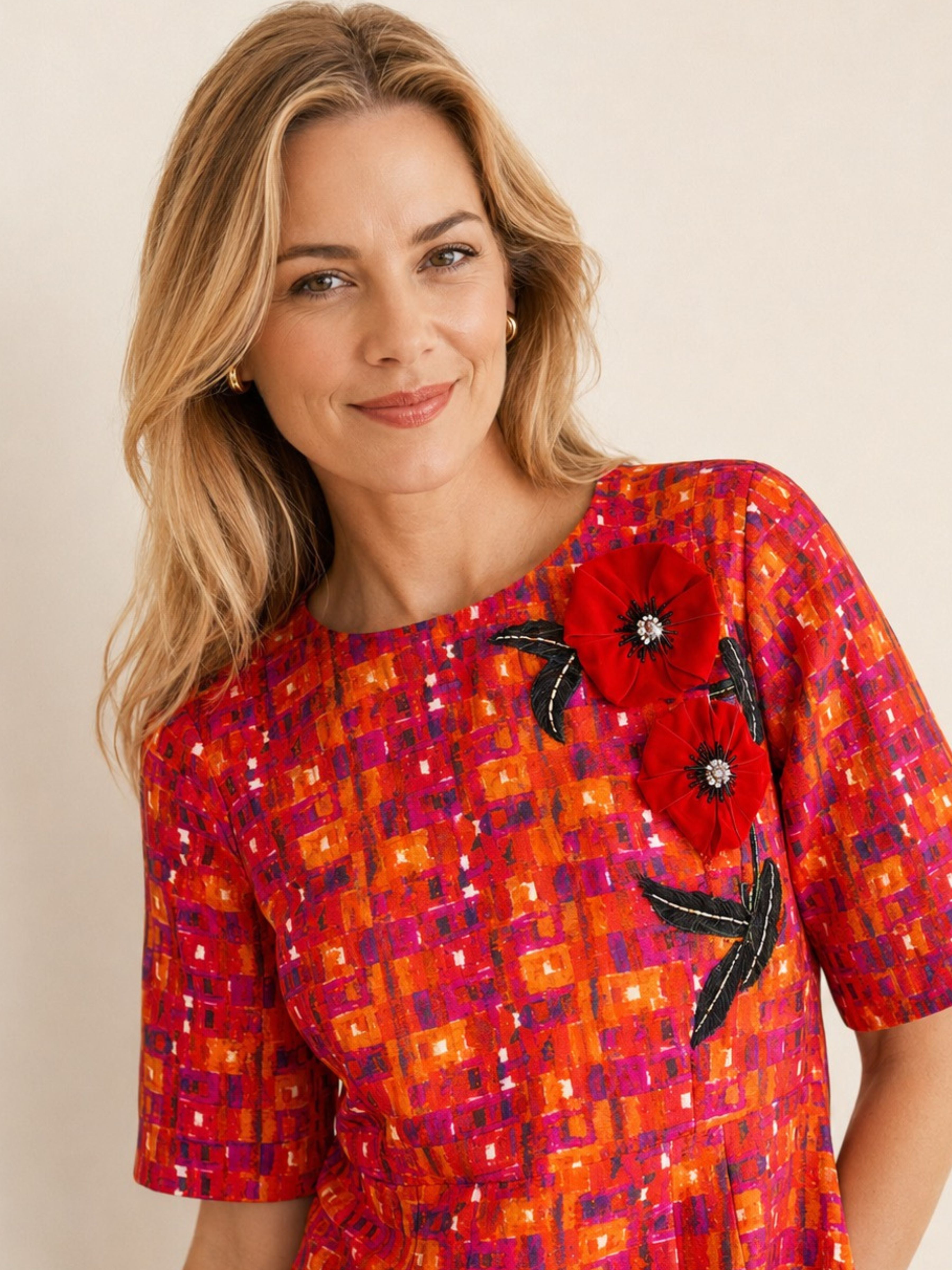 A woman wearing a short-sleeve top with a vibrant orange, red, and magenta geometric pattern, featuring large red poppy appliques with black leaves on the chest.