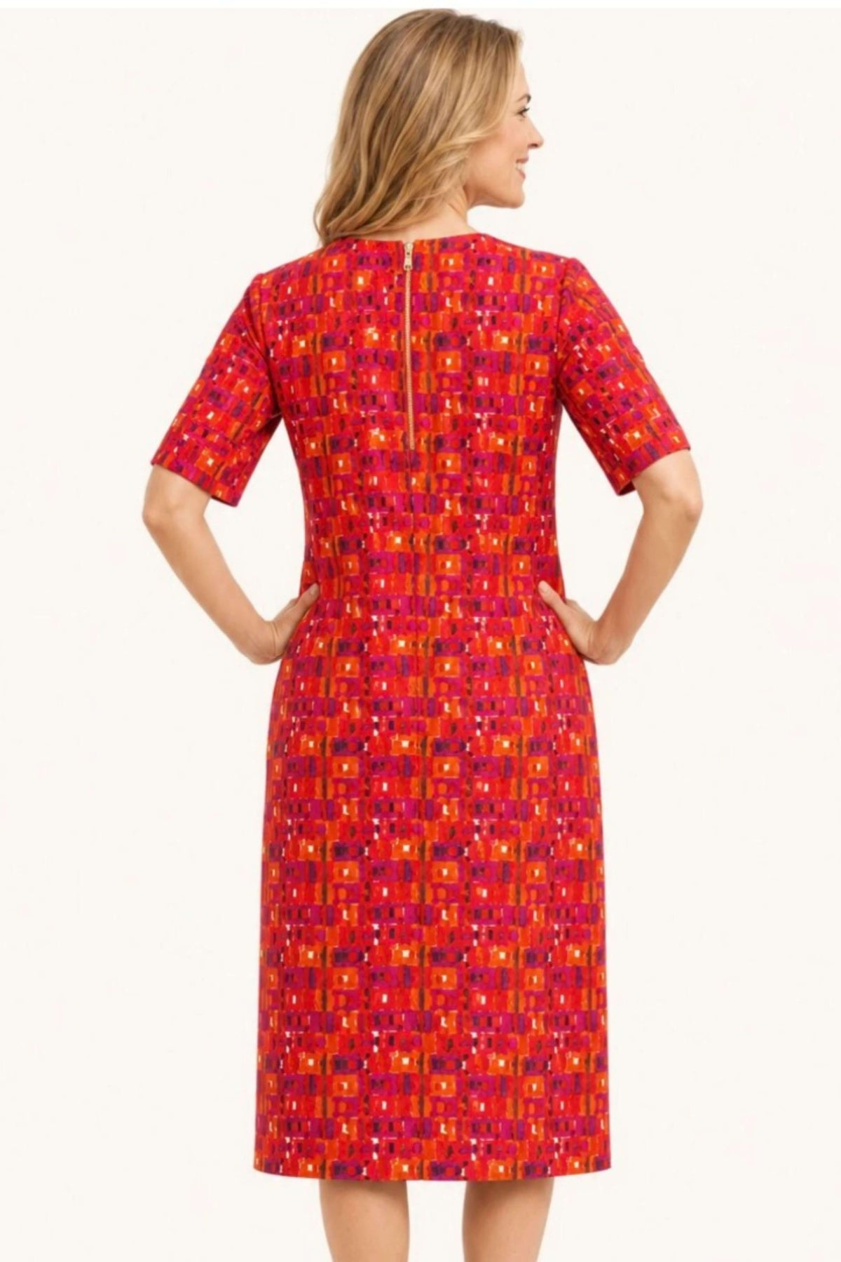 A woman shown from the back wearing a knee-length short-sleeve dress with a vibrant geometric pattern in red, orange, and purple on a plain light background.