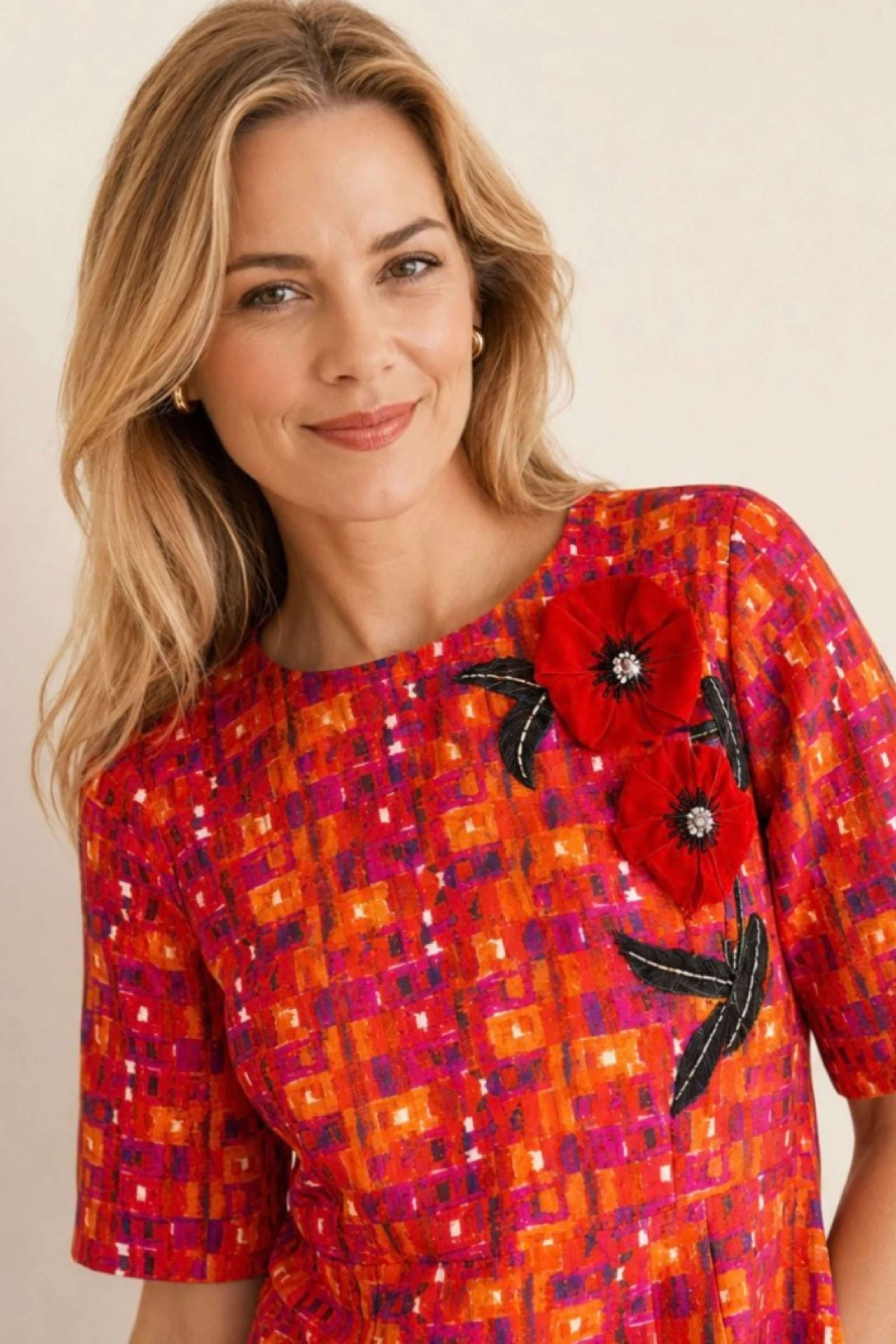 A woman wearing a short-sleeve top with a vibrant orange, red, and magenta geometric pattern, featuring large red embroidered poppy flowers with black leaves on the chest.