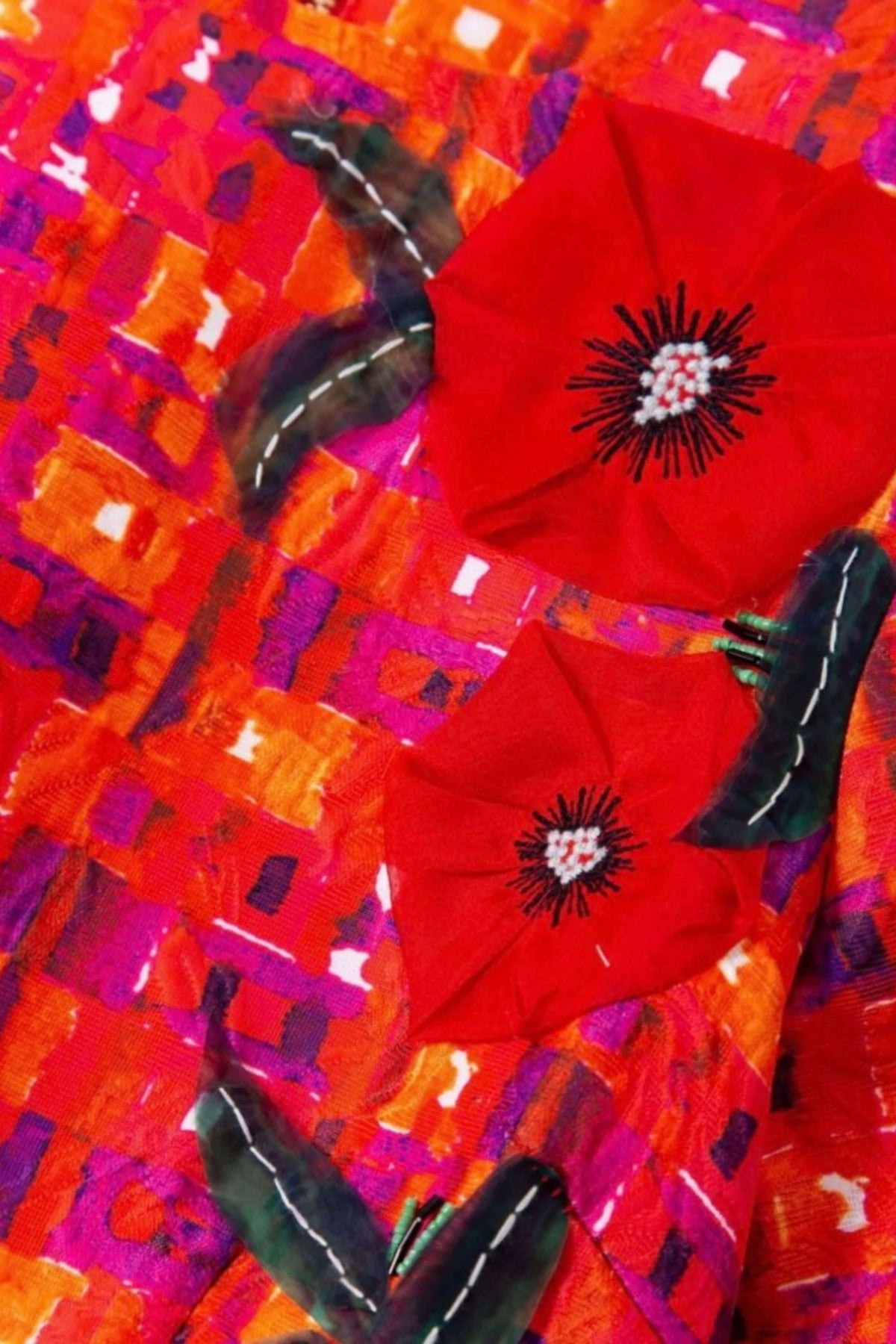 A close-up of a vibrant textile featuring two large red poppy flowers with black and white embroidered centers, set against a highly textured, abstract background of orange, pink, and purple brushstrokes with black feather accents.