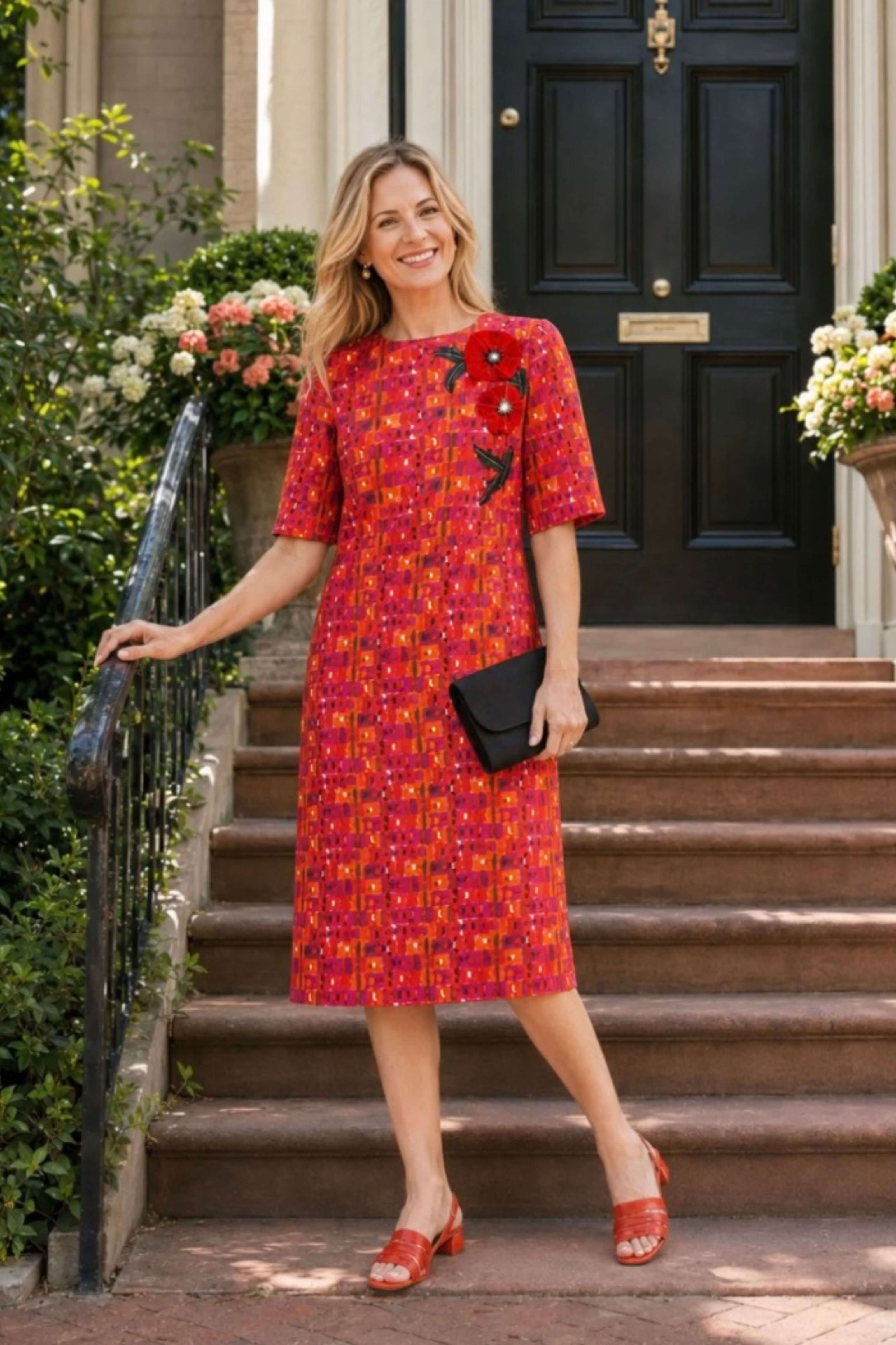 A woman stands on stone steps in front of a building entrance wearing a short-sleeve knee-length dress with an allover red, orange, and yellow floral pattern, plus a black clutch and red sandals.