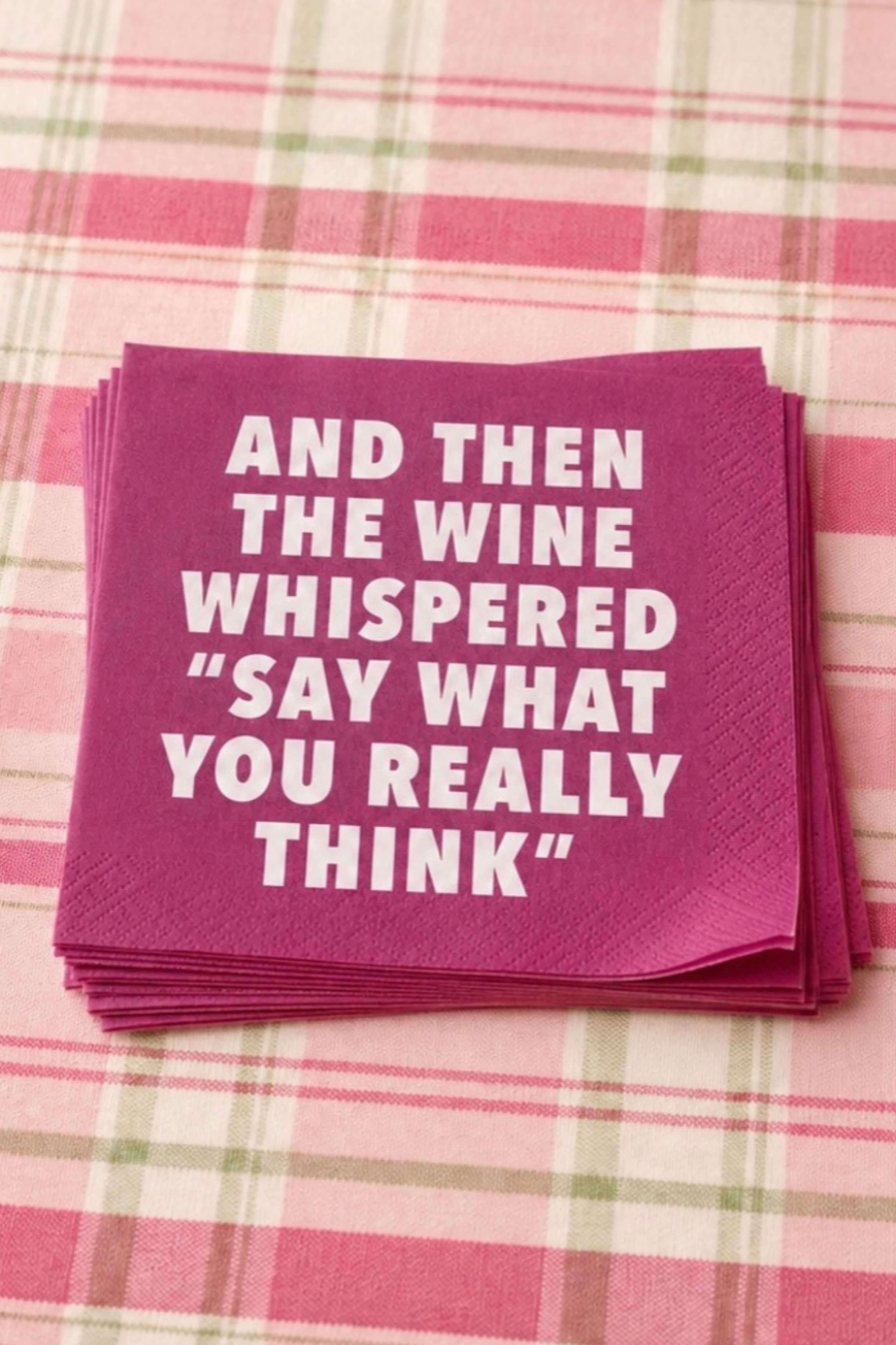 A stack of magenta napkins with bold white text reading 'AND THEN THE WINE WHISPERED "SAY WHAT YOU REALLY THINK"' placed on a pink and green plaid tablecloth.