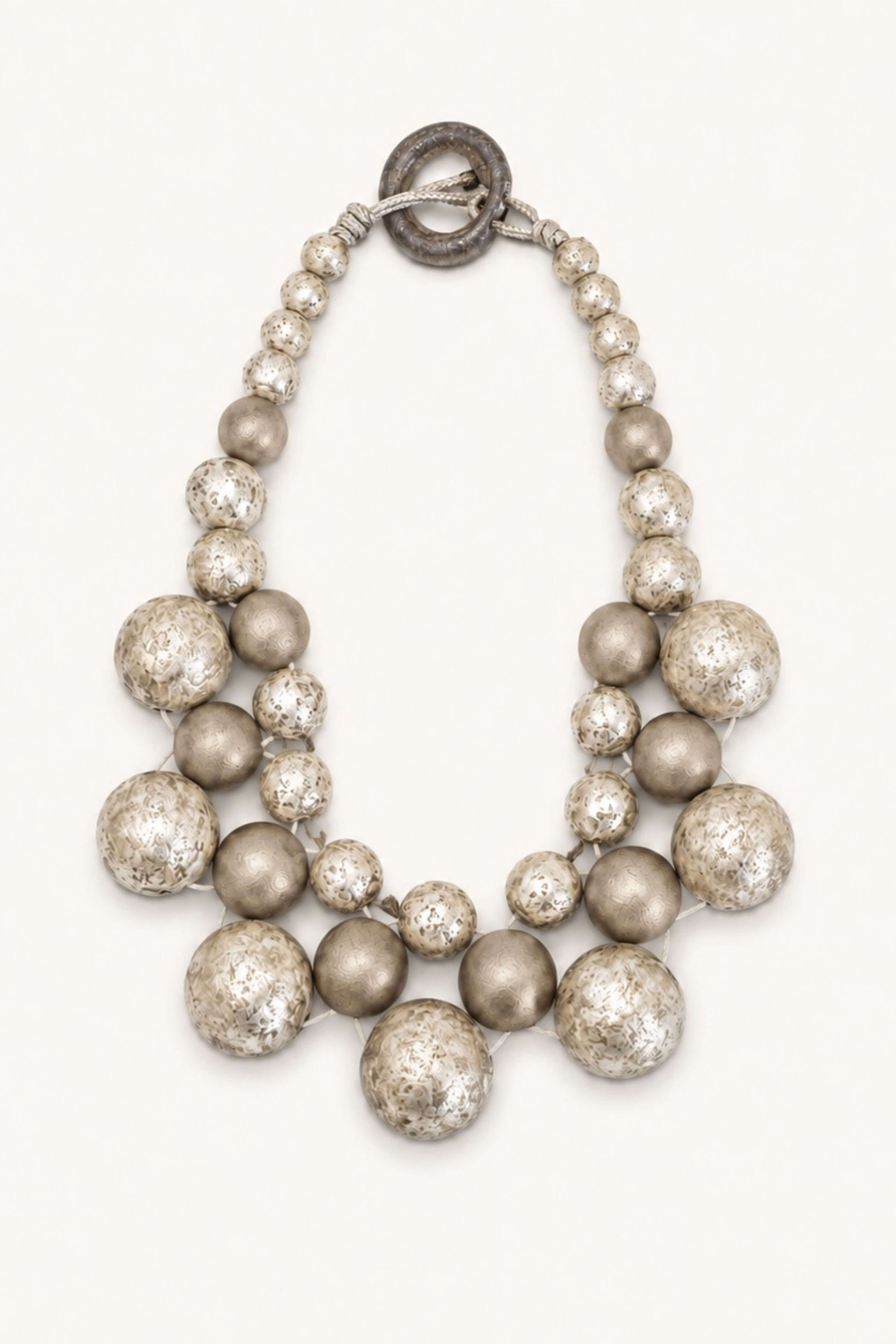 A statement beaded necklace featuring a combination of matte metallic silver, textured metallic silver, and glossy champagne-gold beads, complete with a dark metal toggle clasp, set against a solid off-white background.